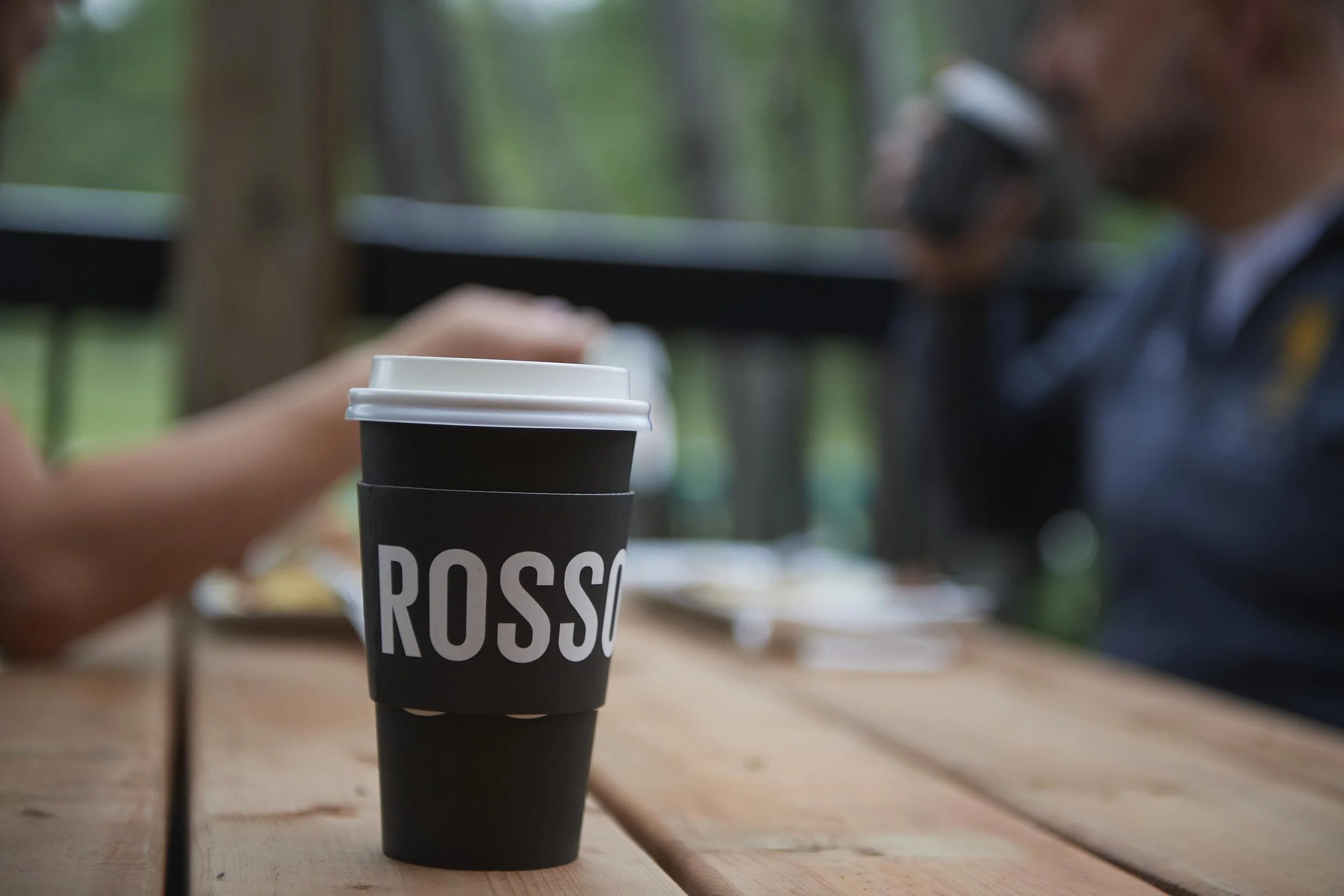 Black coffee cup with a white lid, placed on a wooden table outdoors, with people in the background drinking coffee.