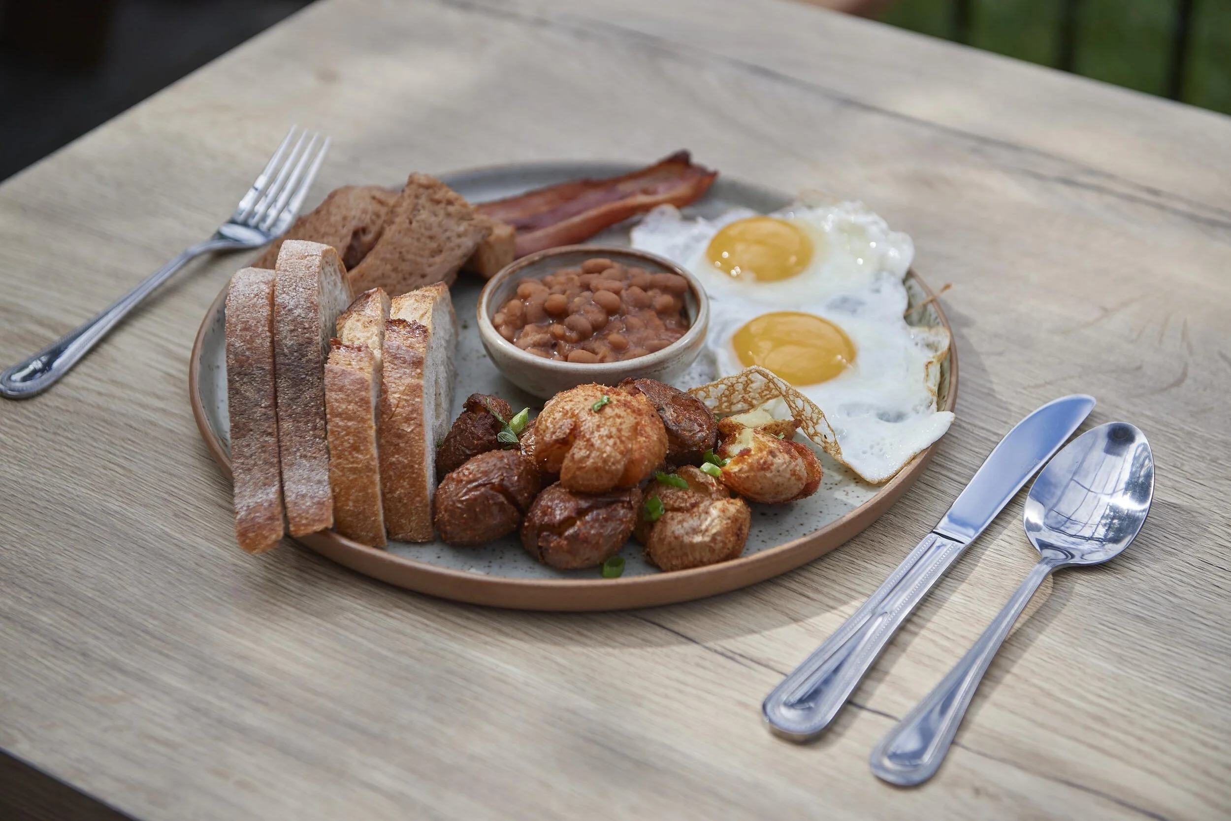Full English breakfast with fried eggs, bacon, baked beans, hash browns, sausage, toast, and a spinach chip, served on a round plate on a wooden table.