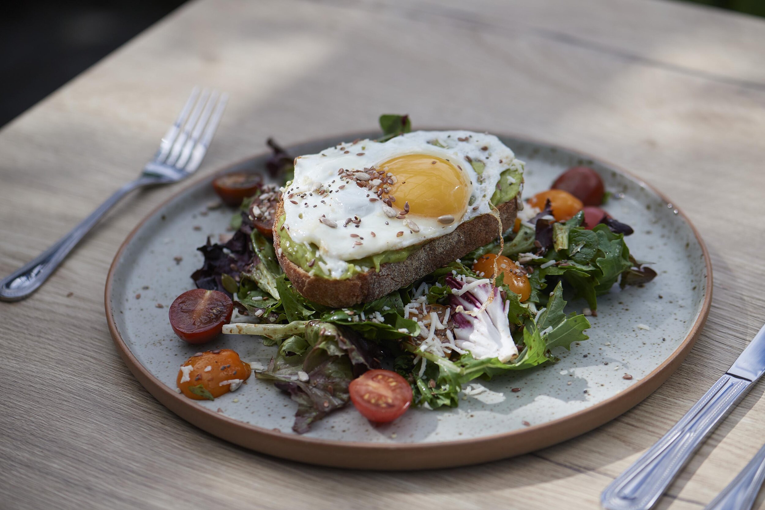 A plate of salad topped with a slice of toasted bread, avocado, a fried egg, and sprinkled seeds, with cherry tomatoes and salad greens around.