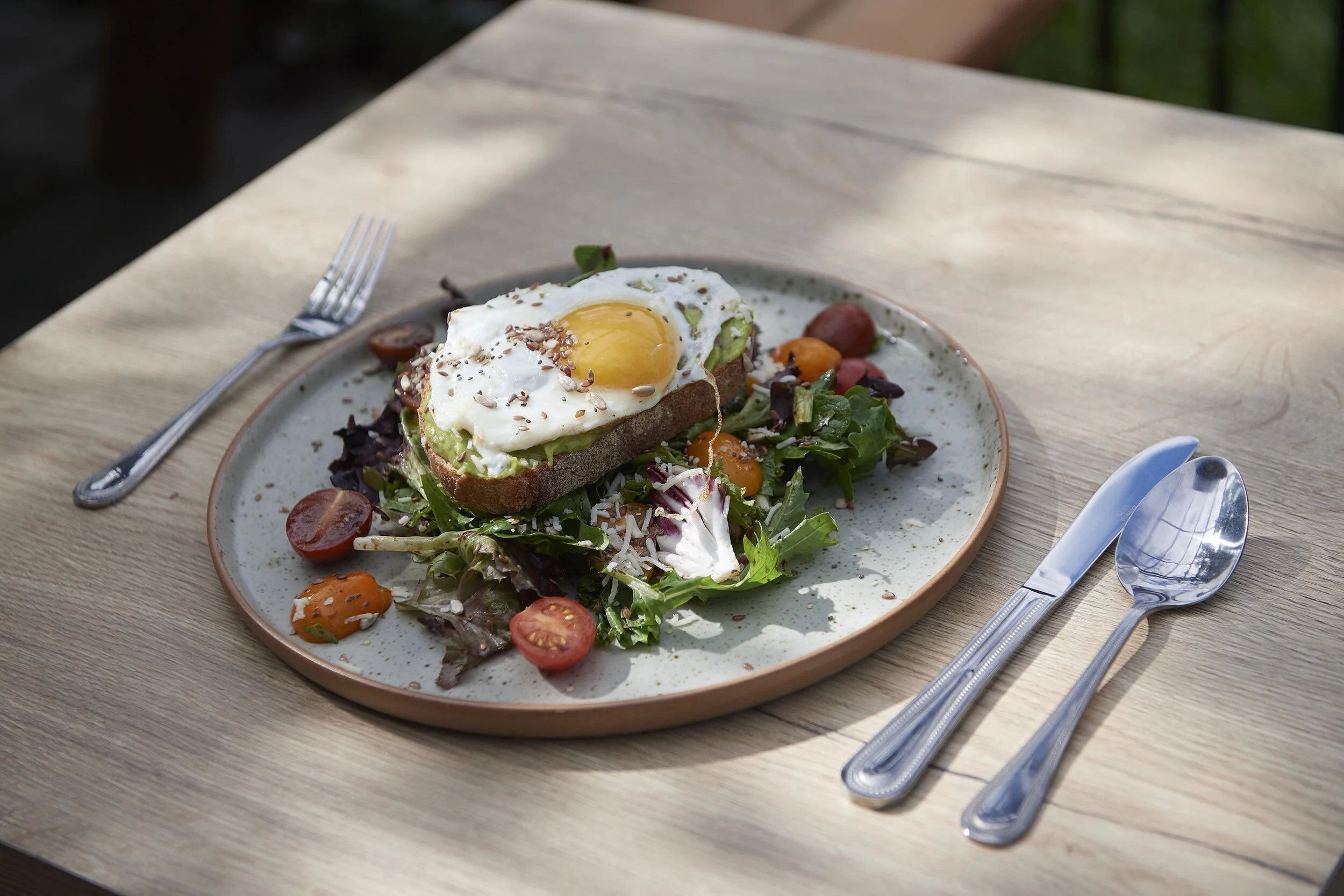 Plate with toast topped with a fried egg and seeds, served over a mixed salad with cherry tomatoes and dressing, on a wooden table with fork, knife, and spoon.