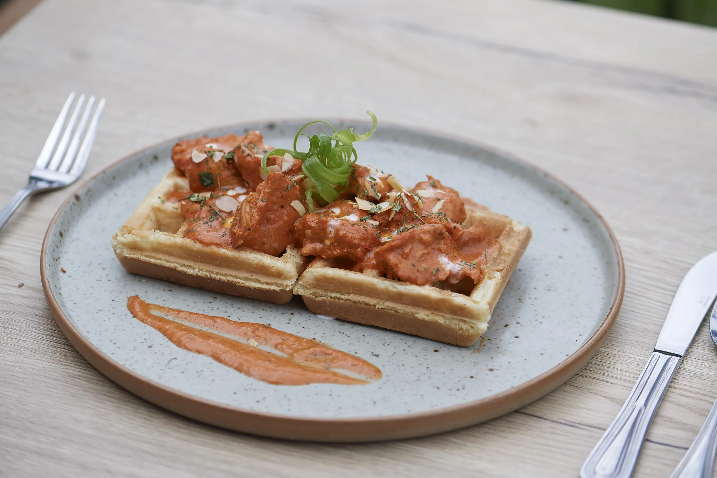 Waffle topped with chicken and sauce, garnished with green onions and herbs, served on a ceramic plate with a drizzle of sauce on the side.
