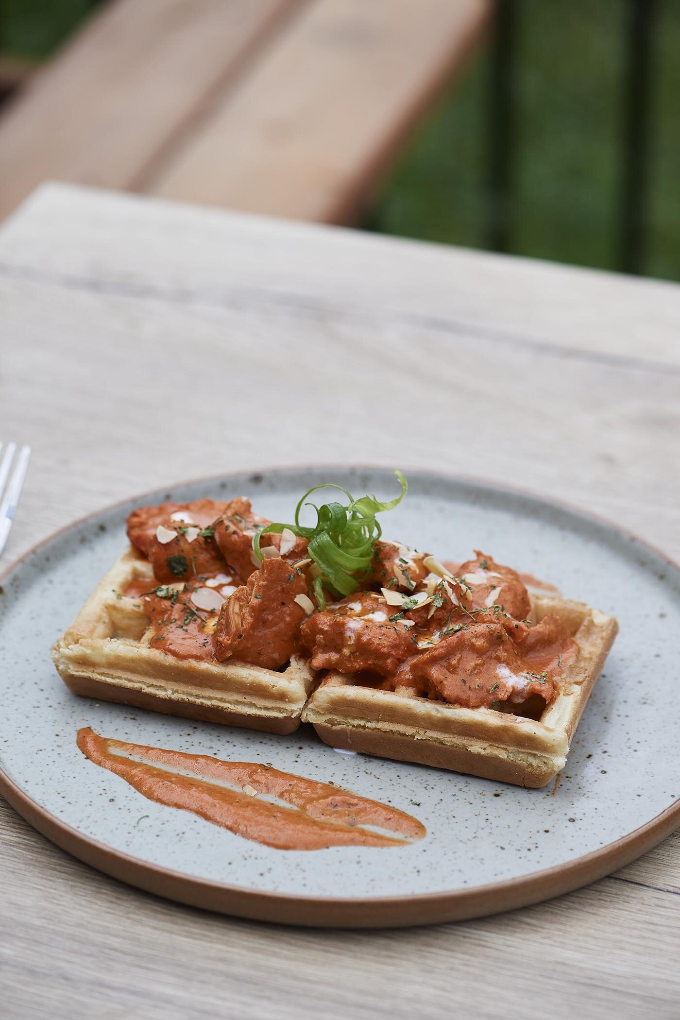 Waffle topped with Butter Chicken, garnished with green onions and sliced almonds, served on a white plate with a smear of sauce.