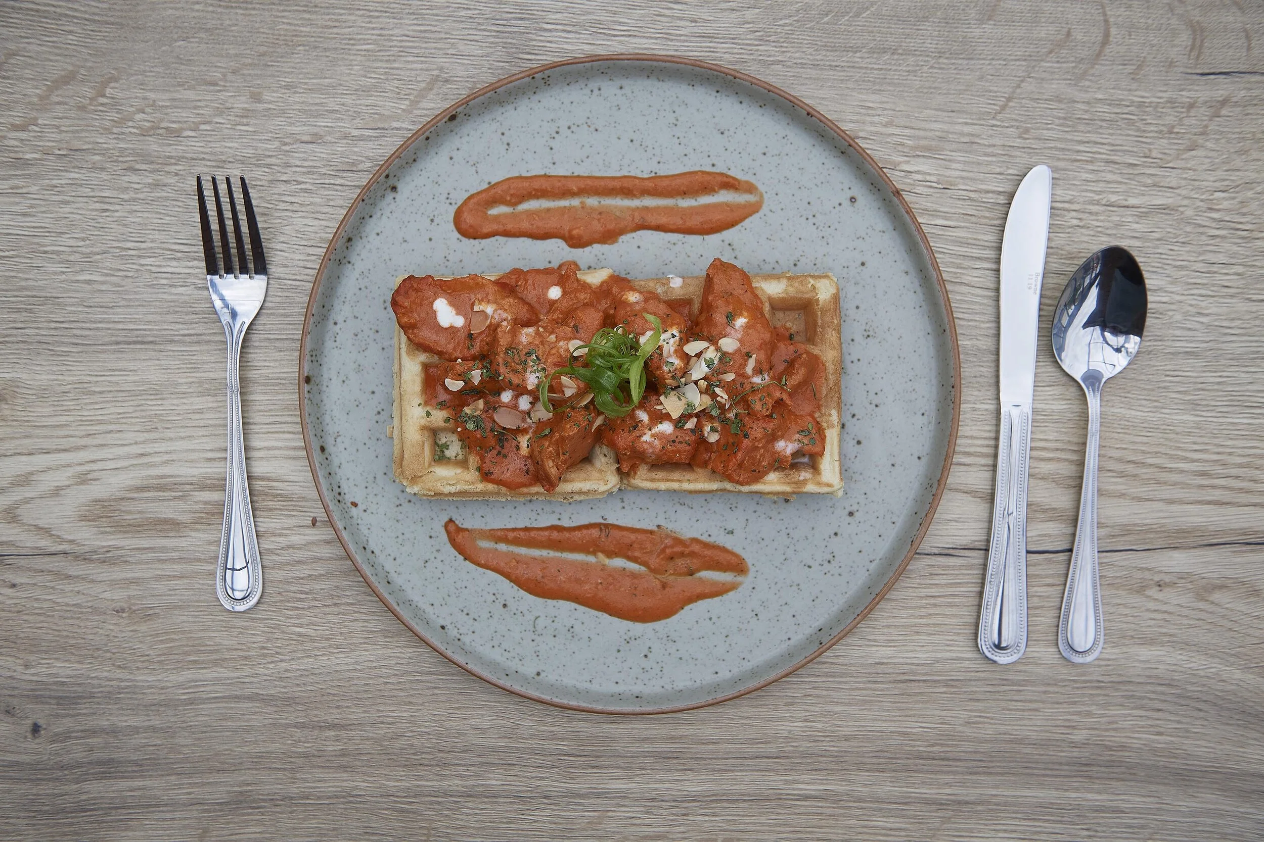 A plate of chicken and waffles topped with sauce and garnished with herbs, placed on a wooden table with a fork on the left, and a knife and spoon on the right.
