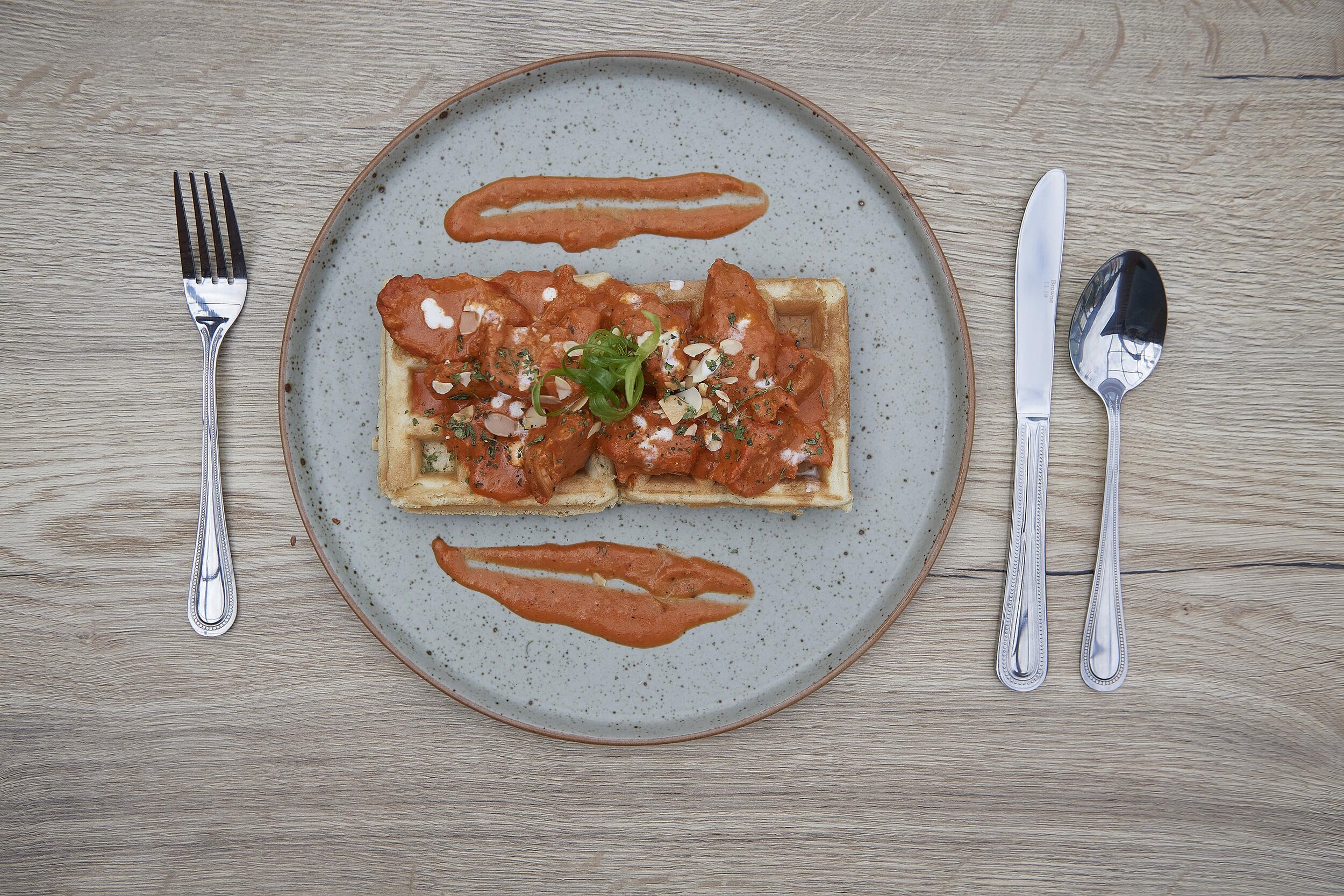 Plate with Butter chicken curry served on a waffle, garnished with green onions and chopped nuts, on a speckled gray plate. There are two dollops of sauce on the plate, one near the top and the other near the bottom. 