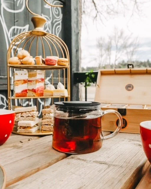 A glass teapot with tea, a layered cake stand with assorted pastries and macarons, a tray with sandwiches, and red bowls on a wooden table outside with trees and sky in the background.