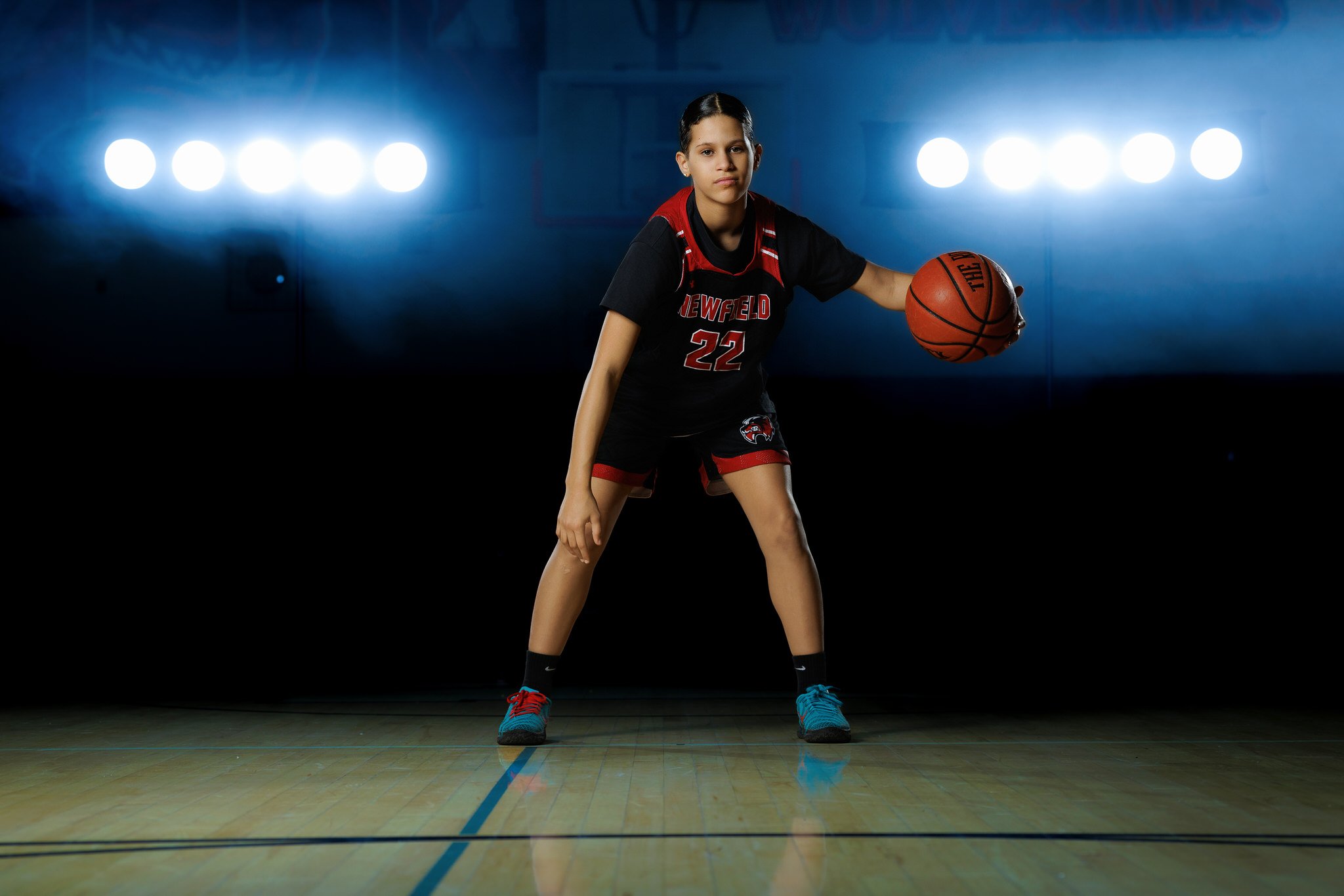 A young female basketball player in a black and red uniform with the number 22, holding a basketball on a court with bright lights in the background.