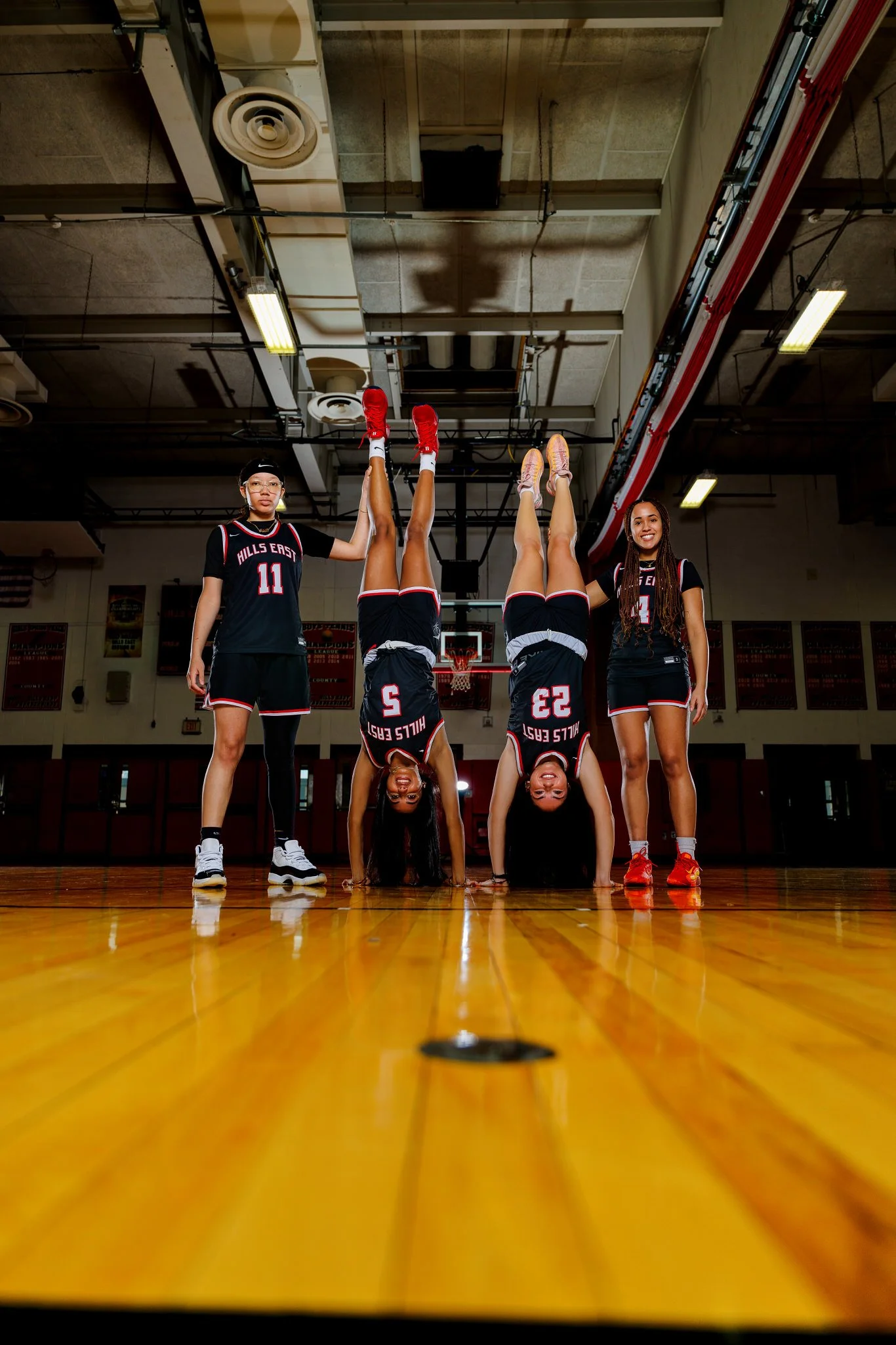 Four female basketball players in black uniforms on a gymnasium court: two are doing handstands, while two stand on either side holding their legs. All are smiling.