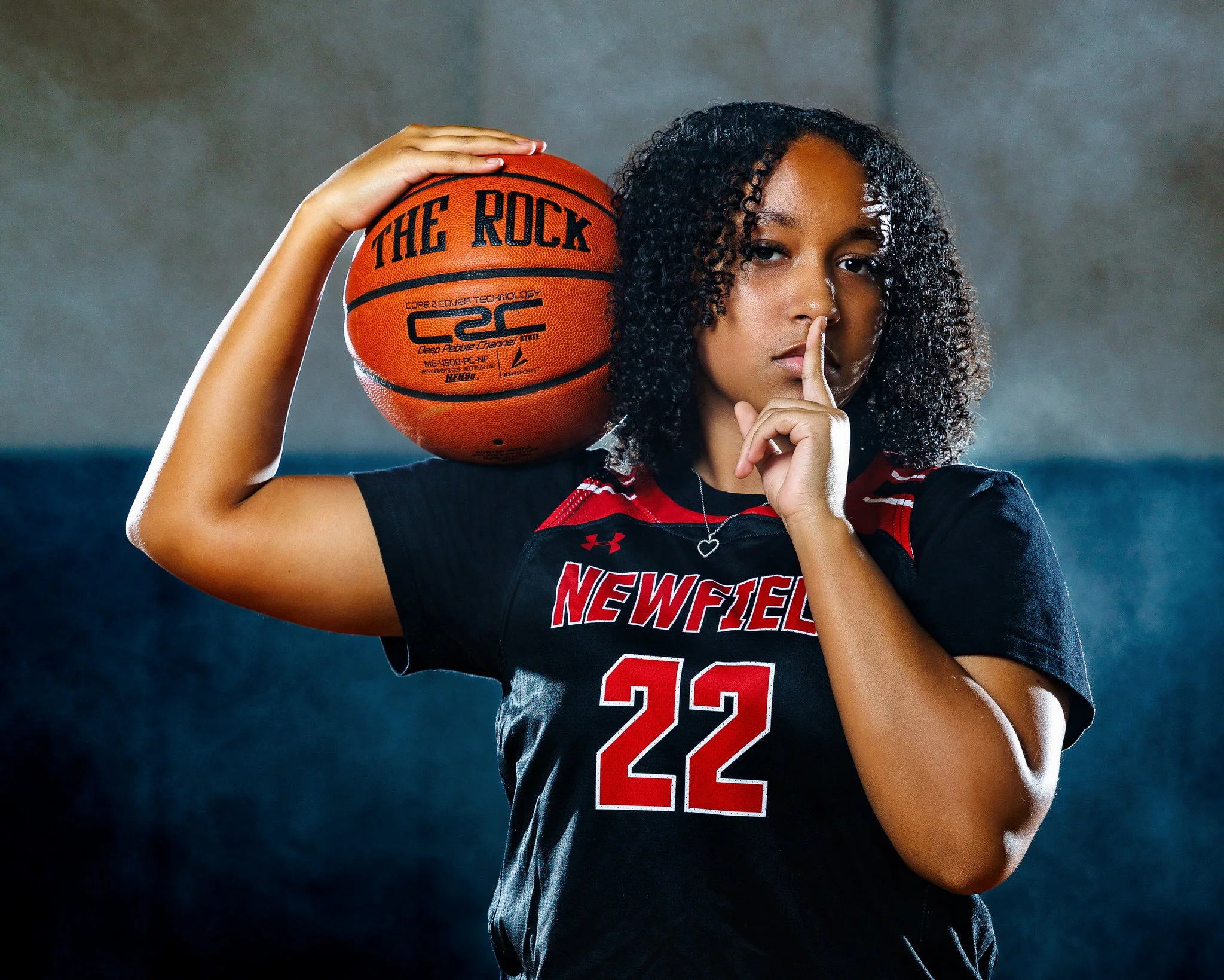 A female basketball player in a black and red jersey with the number 22, holding a basketball on her shoulder and making a finger to lips gesture.