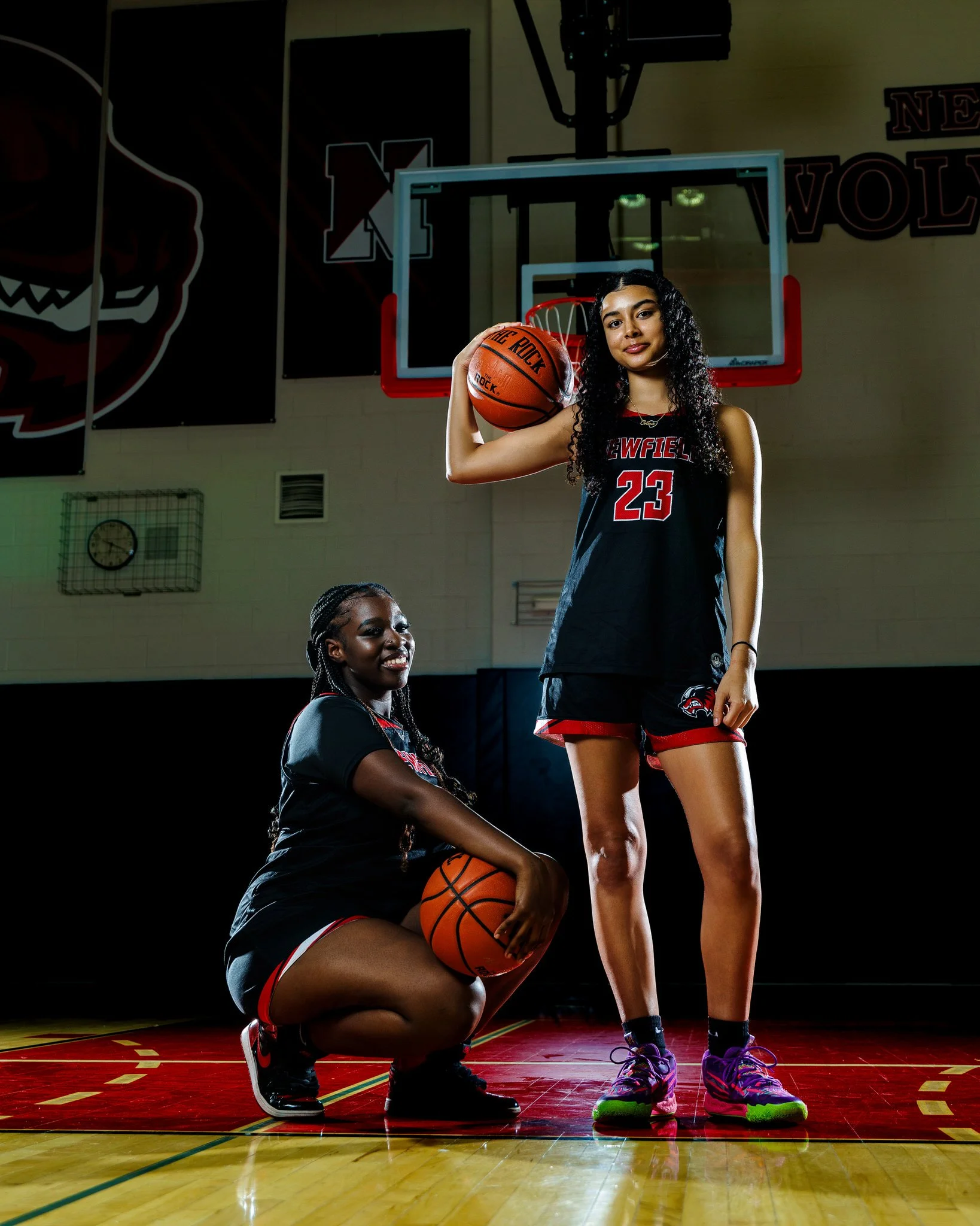 Two female basketball players in athletic uniforms on a basketball court in a gym, one standing with a basketball resting on her shoulder, the other crouching holding a basketball, with a basketball hoop and gymnasium decor in the background.