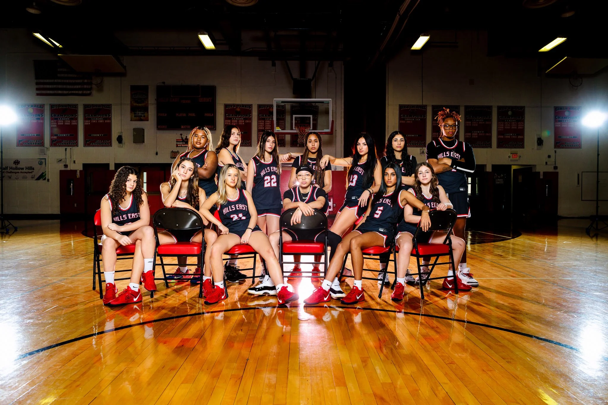 A girls' basketball team posing on a gymnasium basketball court. The team members are wearing black jerseys with red and white accents, with the text 'Hills East' and various numbers, and red basketball shoes. Some players are sitting on chairs while