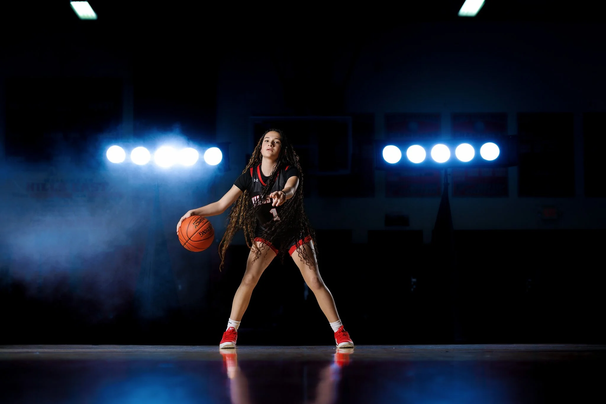 Young girl in a basketball uniform dribbling a basketball on a basketball court, with bright lights and a dark background.