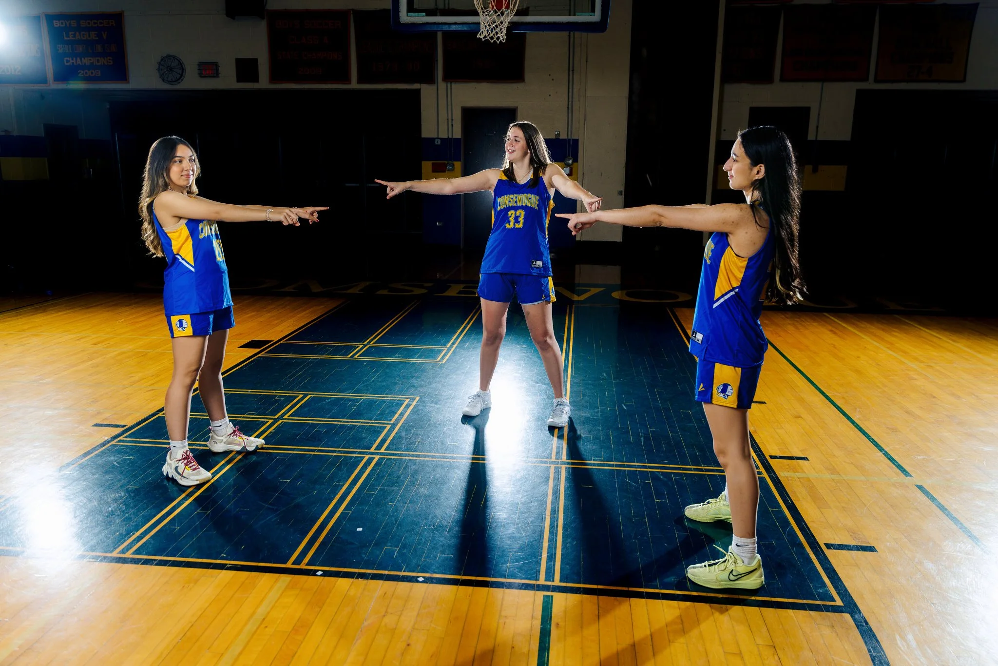 Three young women in blue and yellow basketball uniforms standing on a basketball court, pointing at each other with extended arms, indoors at a gymnasium.