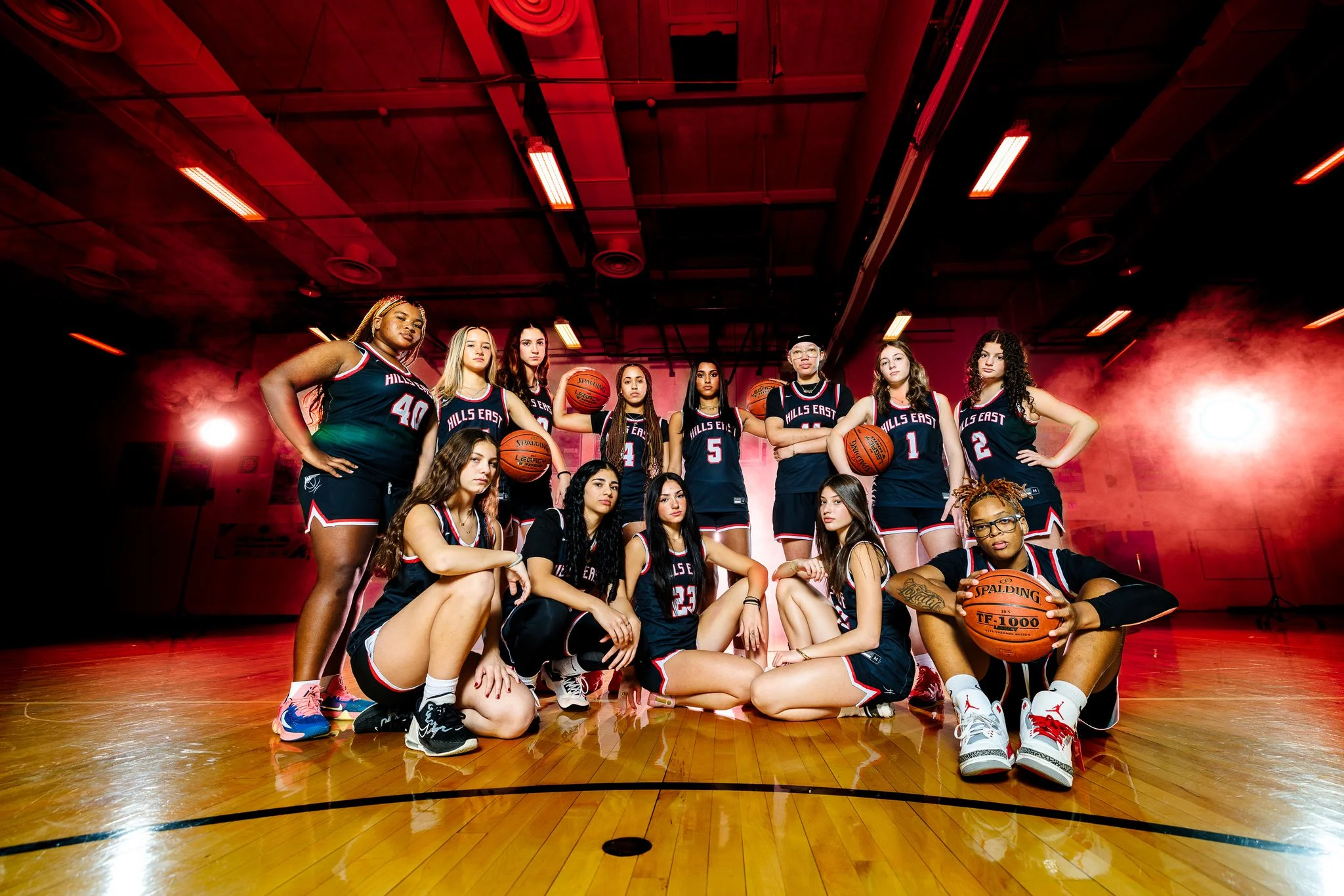 A girls' basketball team posing on a gymnasium court. The team members are wearing black jerseys with red and white accents, and some are holding basketballs. The gym has a wooden floor, red lighting effects, and a high ceiling.