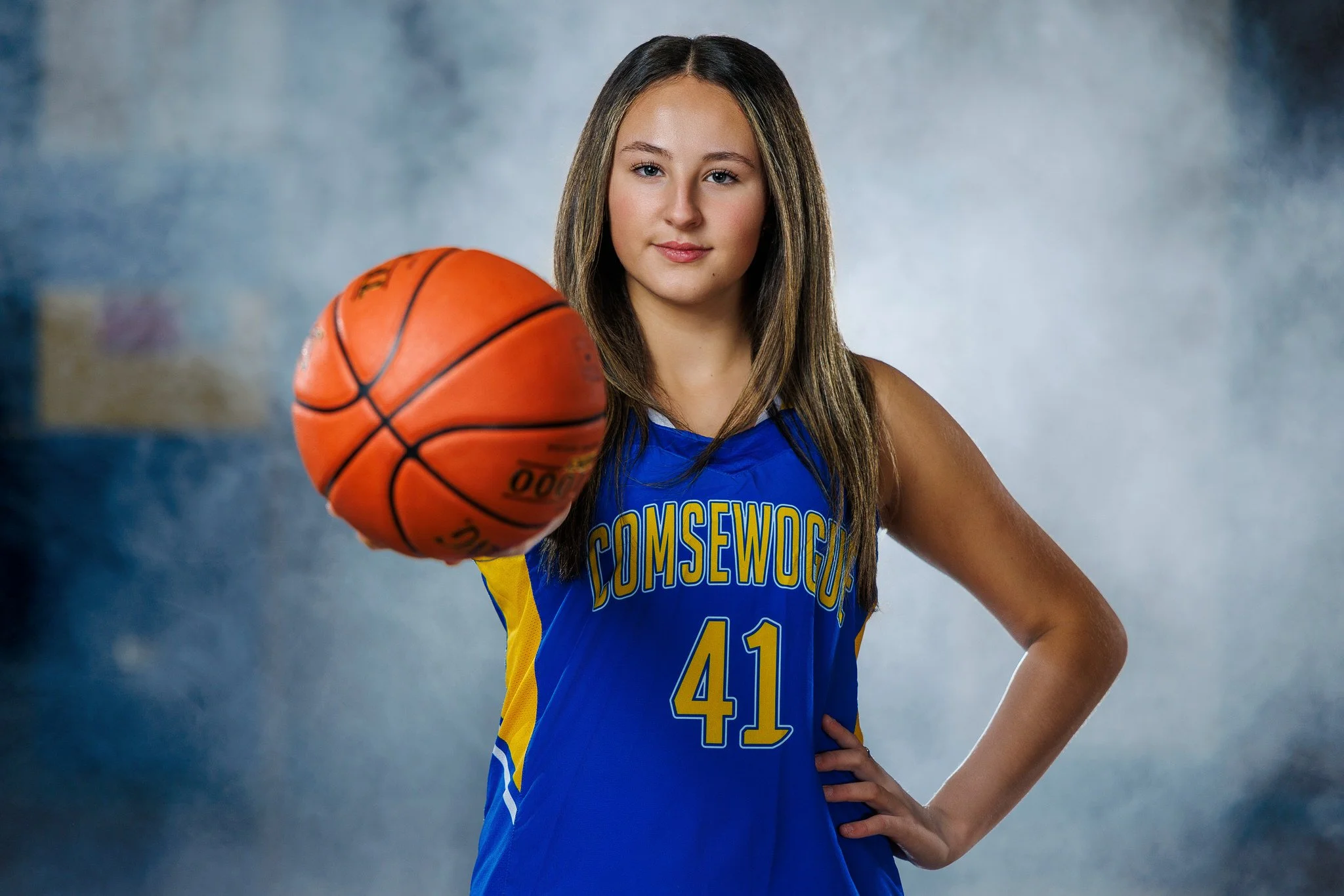 A young female basketball player wearing a blue jersey with yellow accents, number 41, and 'COMSEWOG' written on it, holding a basketball in front of her, standing with her right hand on her hip against a smoky background.