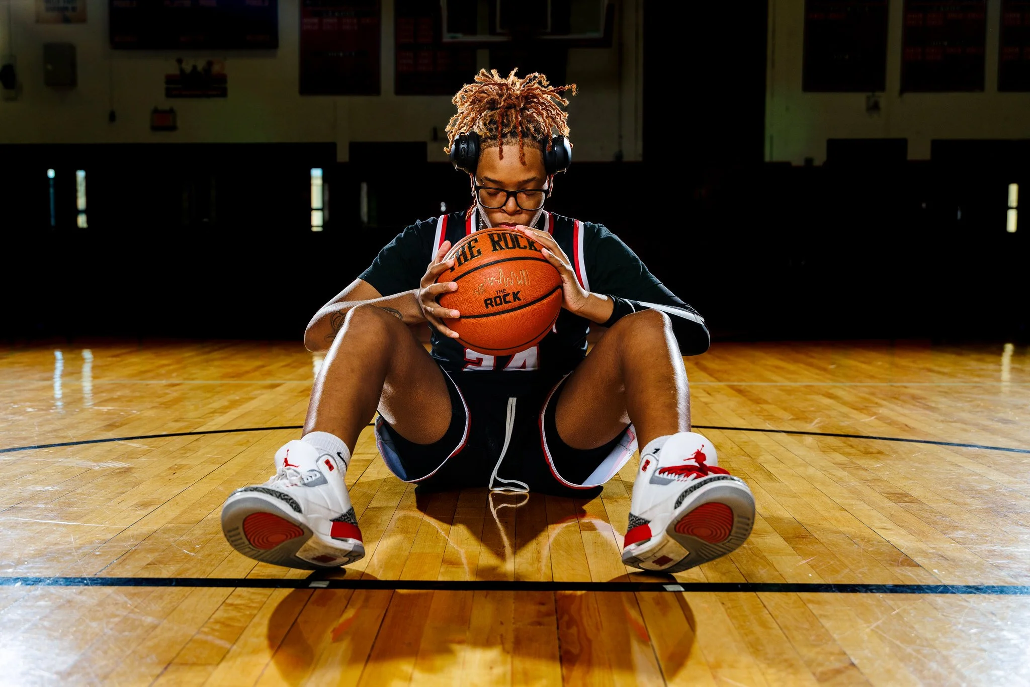 A young woman with glasses and dreadlocks is sitting on a basketball court with her legs spread apart. She is holding a basketball with both hands, sitting on the floor, and wearing a sports jersey, shorts, white and red sneakers, and headphones. The
