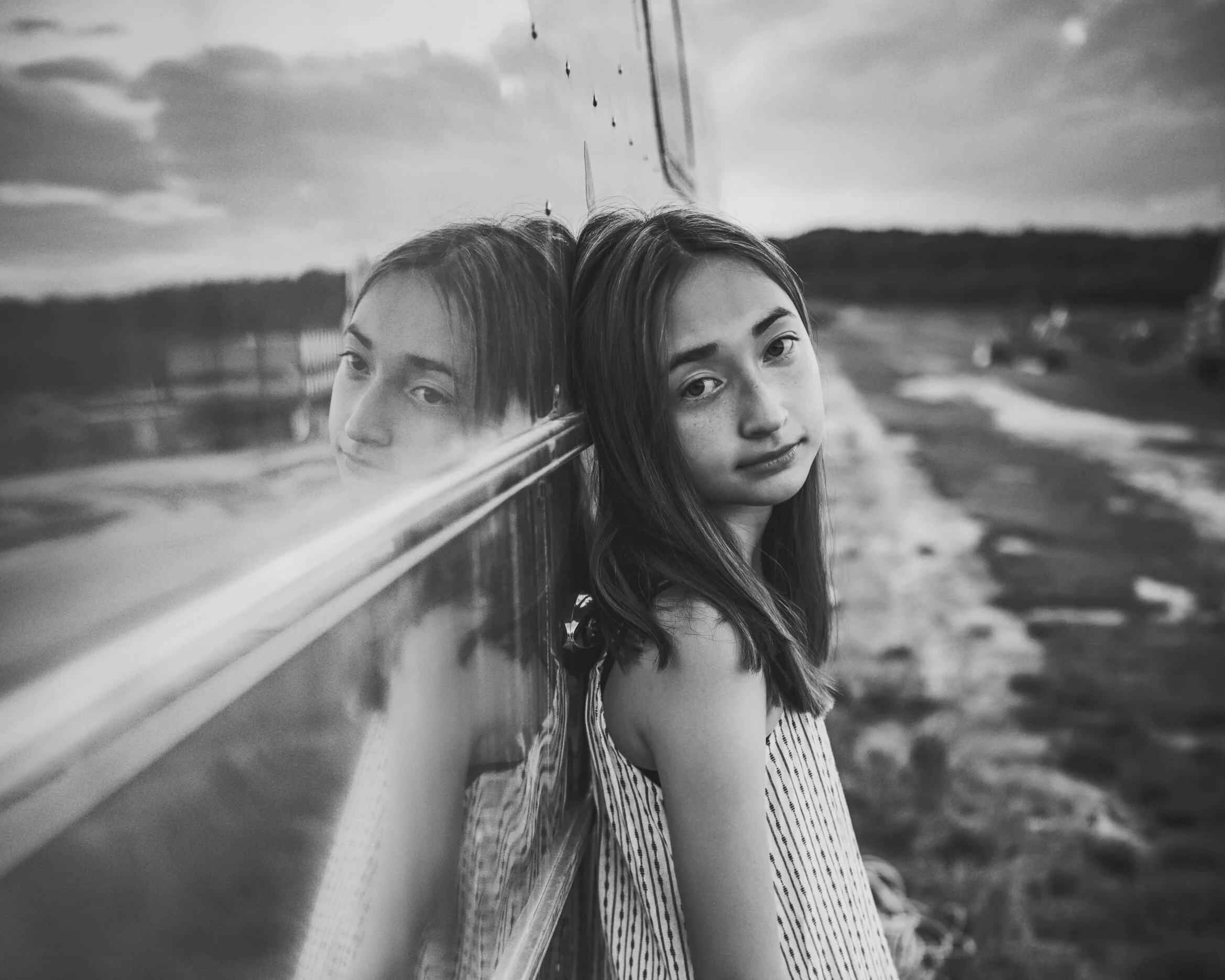 A black and white photo of Megan with long hair leaning against a reflective surface, such as a window or mirror, outdoors with cloudy sky and landscape in background.