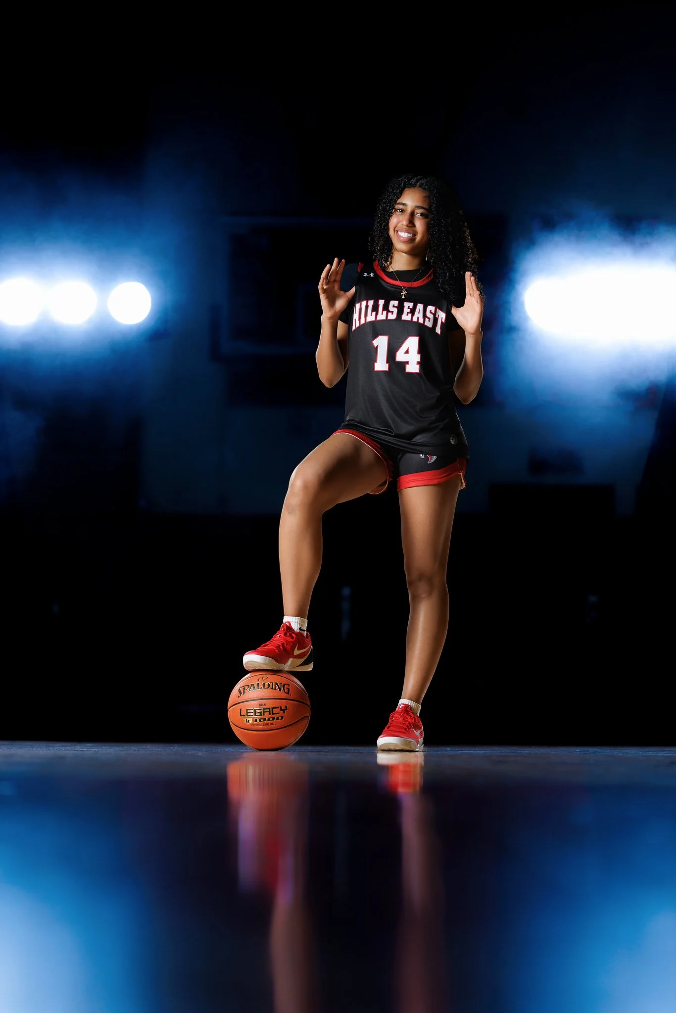 A female basketball player stands on a basketball court with one foot on a basketball. She is smiling, wearing a black and red jersey with the number 14 and the words 'Hills East' on it, and red sneakers. The background is dark with four bright light