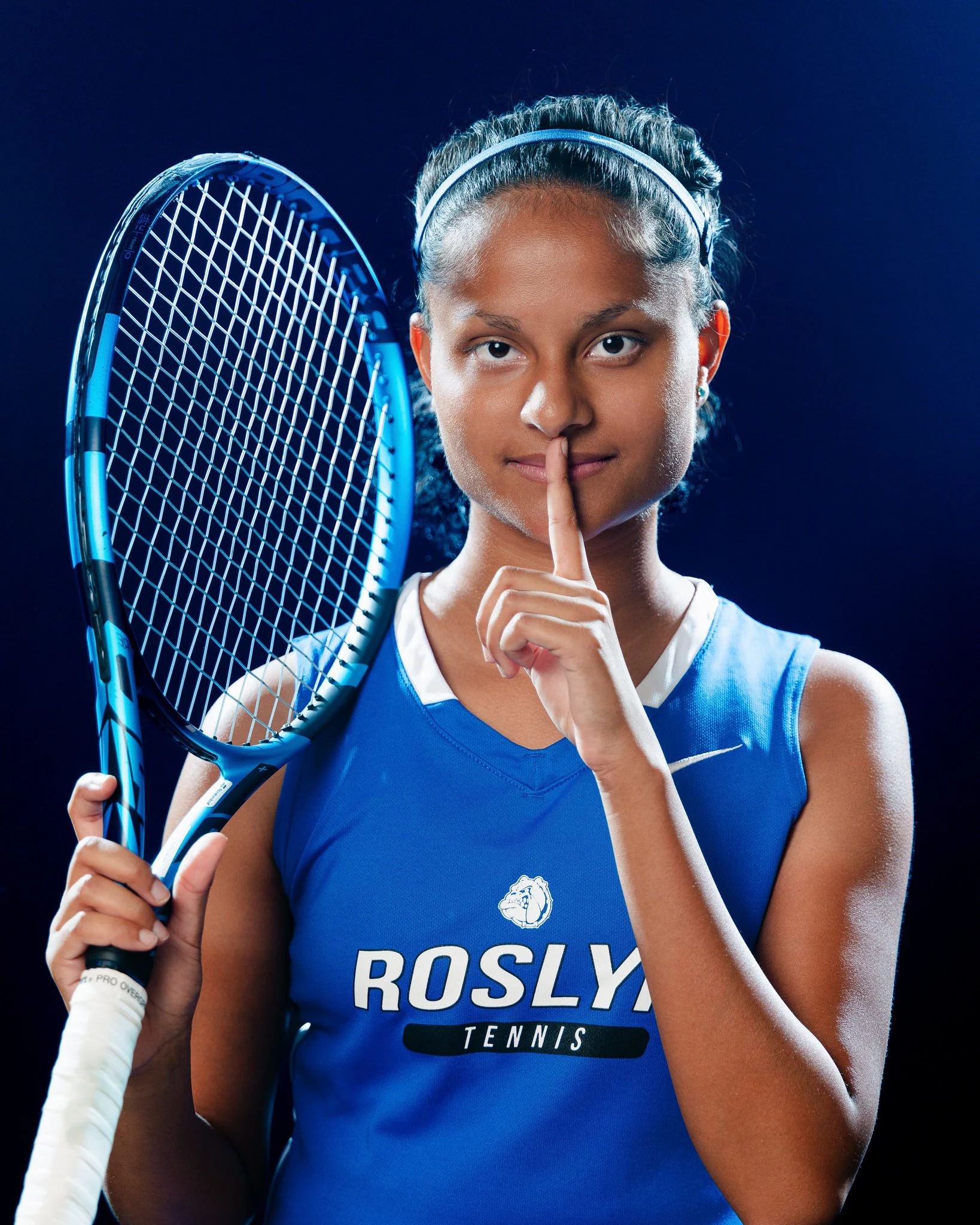 A young female tennis player holding a tennis racket, wearing a blue sports jersey and making a 'shh' gesture with her finger to her lips.