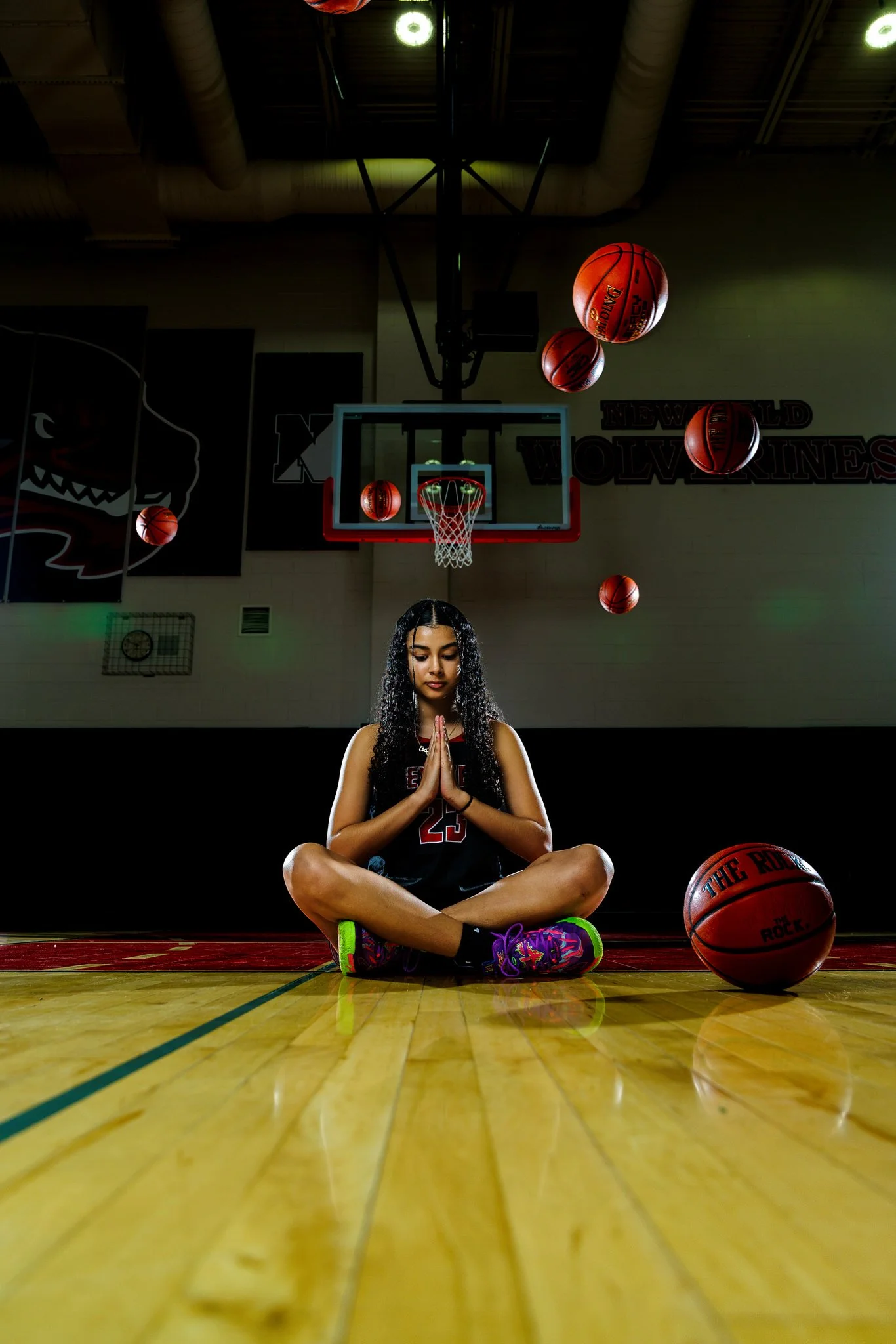 A young girl sitting cross-legged on a basketball court with her hands in a prayer pose, surrounded by floating basketballs, in a gymnasium with a basketball hoop and team banners in the background.