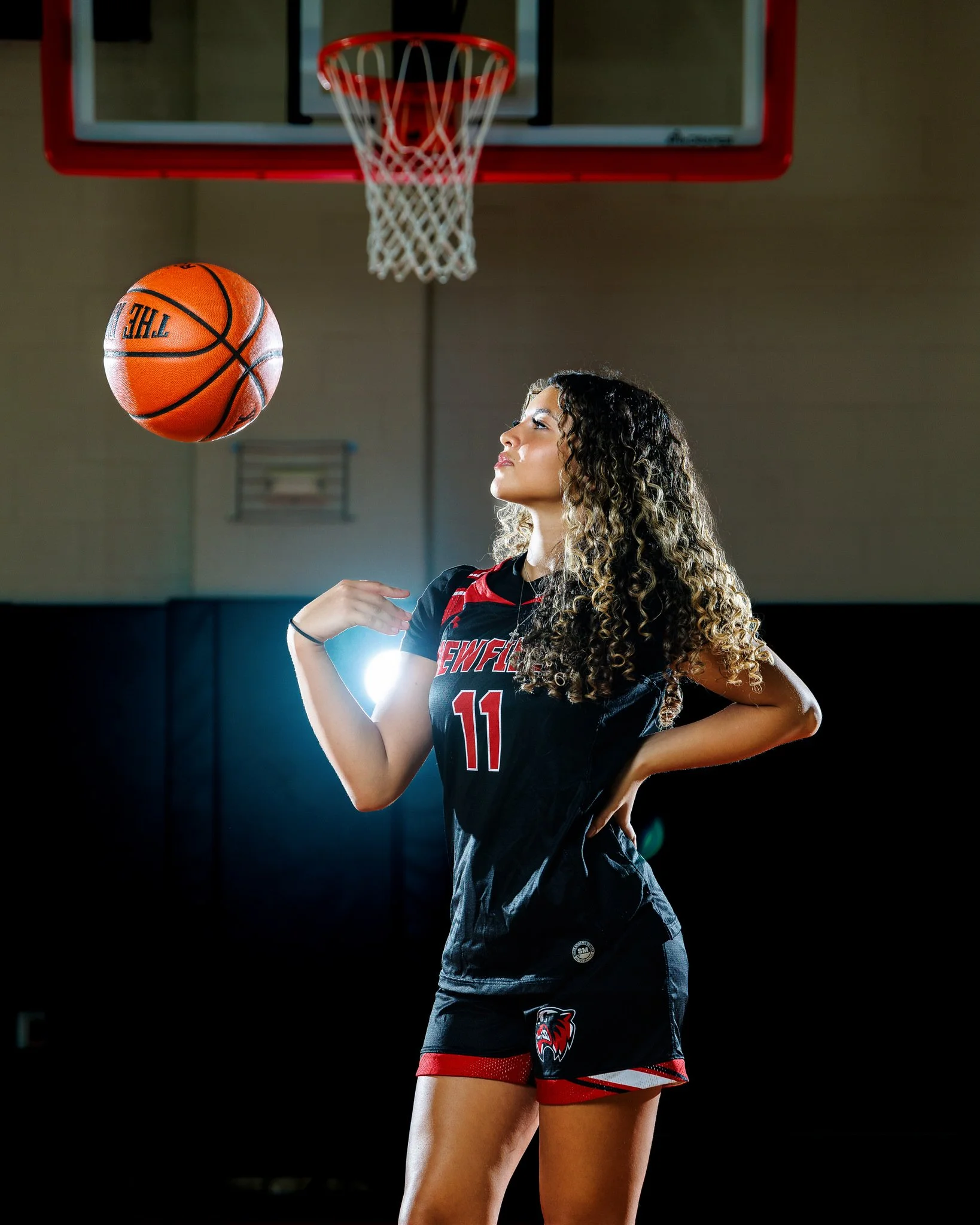 A female volleyball player in a black and red uniform standing in a gymnasium, looking at a basketball mid-air near the basketball hoop.