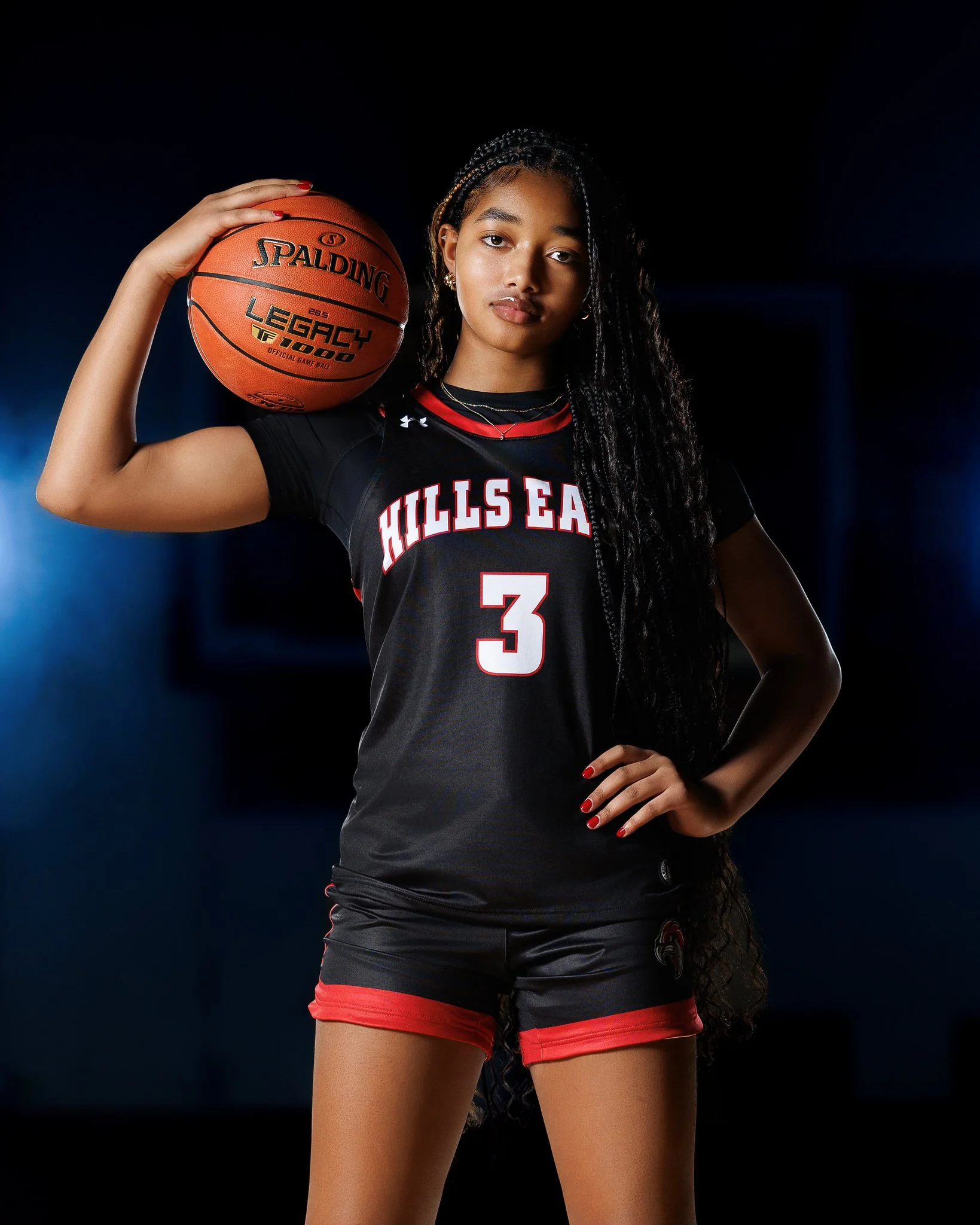 A young woman holding a basketball on her shoulder, wearing a black and red basketball uniform with the number 3, in a dimly lit gym.