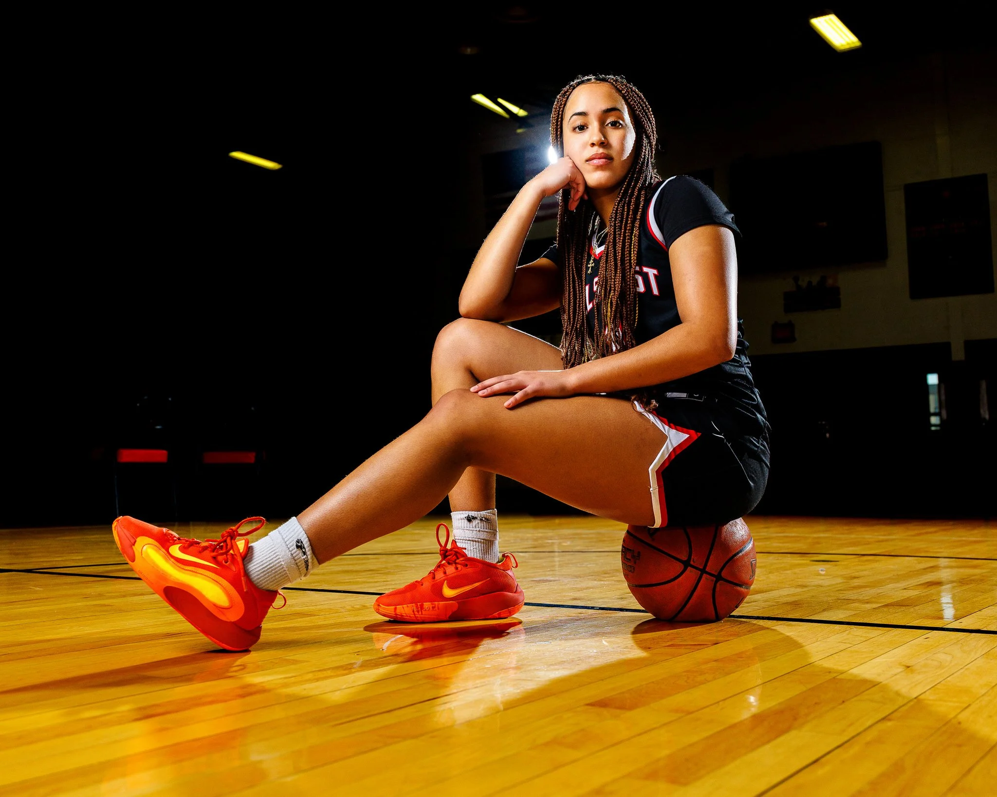 Young woman in a black basketball uniform with red seaming, sitting on a basketball on an indoor court with hardwood flooring, illuminated by overhead lights, with a basketball resting under her. She wears bright red Nike sneakers, white socks, and h