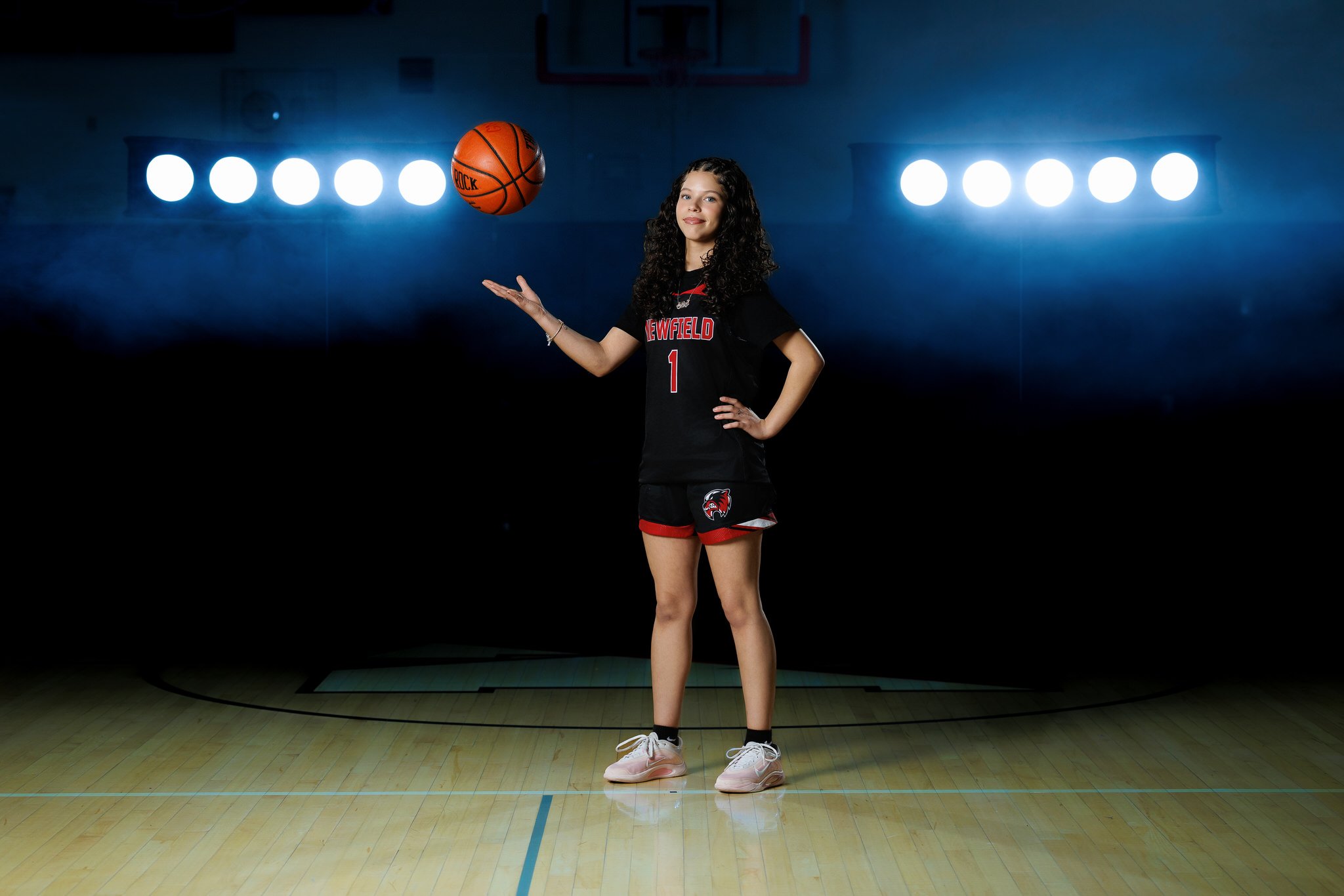 A young woman with curly hair in a black and red basketball uniform spinning a basketball on her finger on a basketball court.