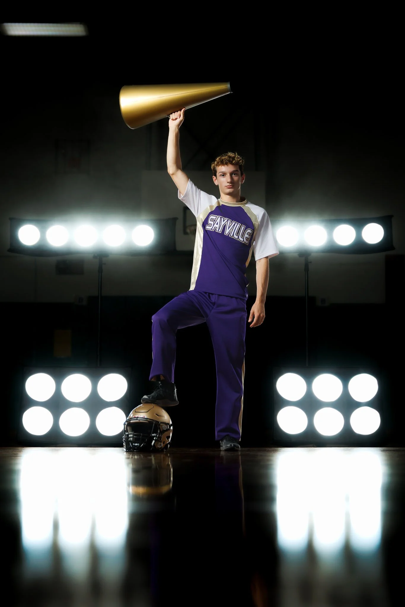 A young male volleyball player in a purple and white jersey with 'Sayville' on it, standing on a volleyball court with one foot on a helmet and the other on a volleyball, holding a golden megaphone above his head, with bright stage lights in the back