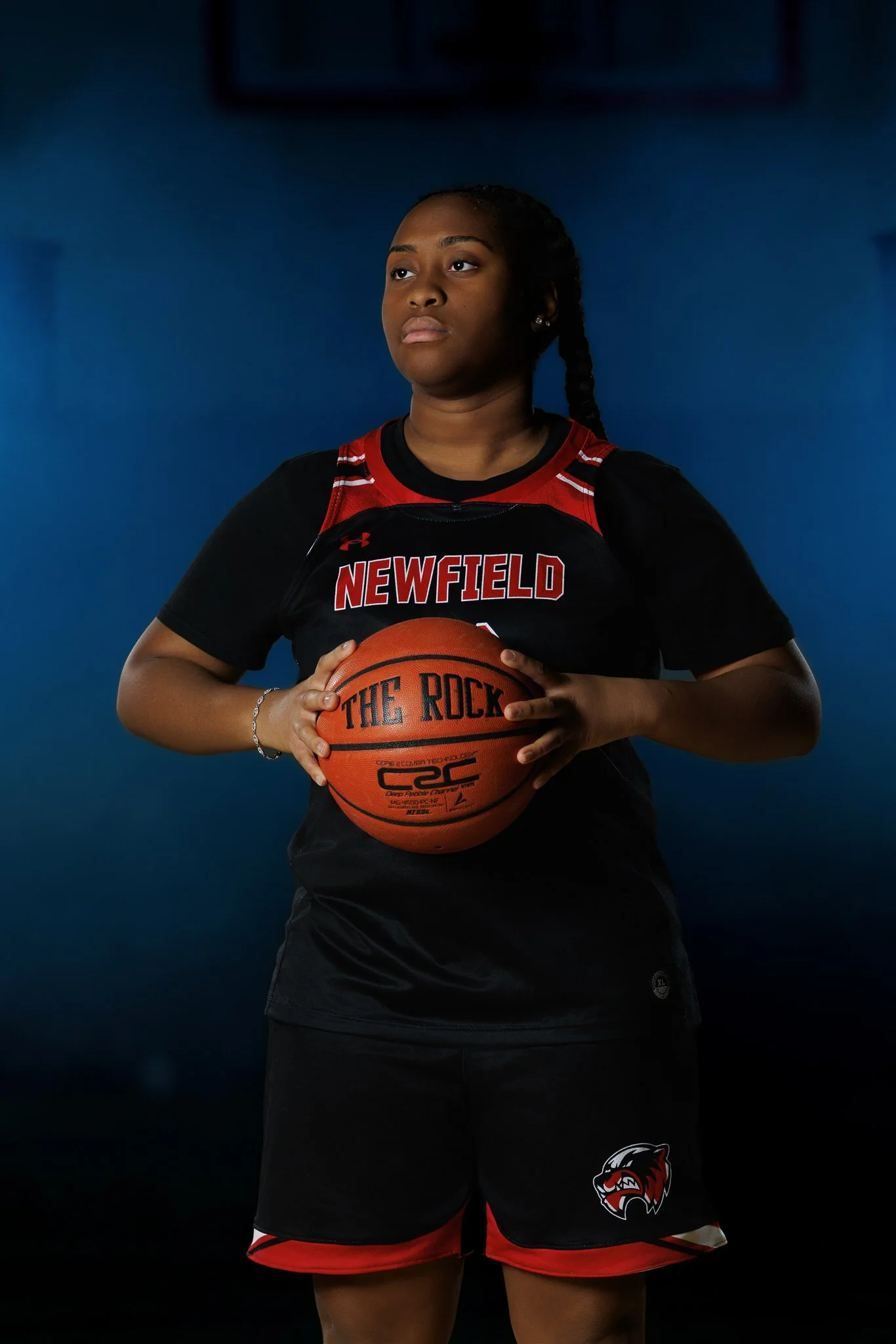A female basketball player standing against a dark blue background, holding a basketball, wearing a black and red Newfield jersey with 'The Rock' written on it, and shorts with a team logo.