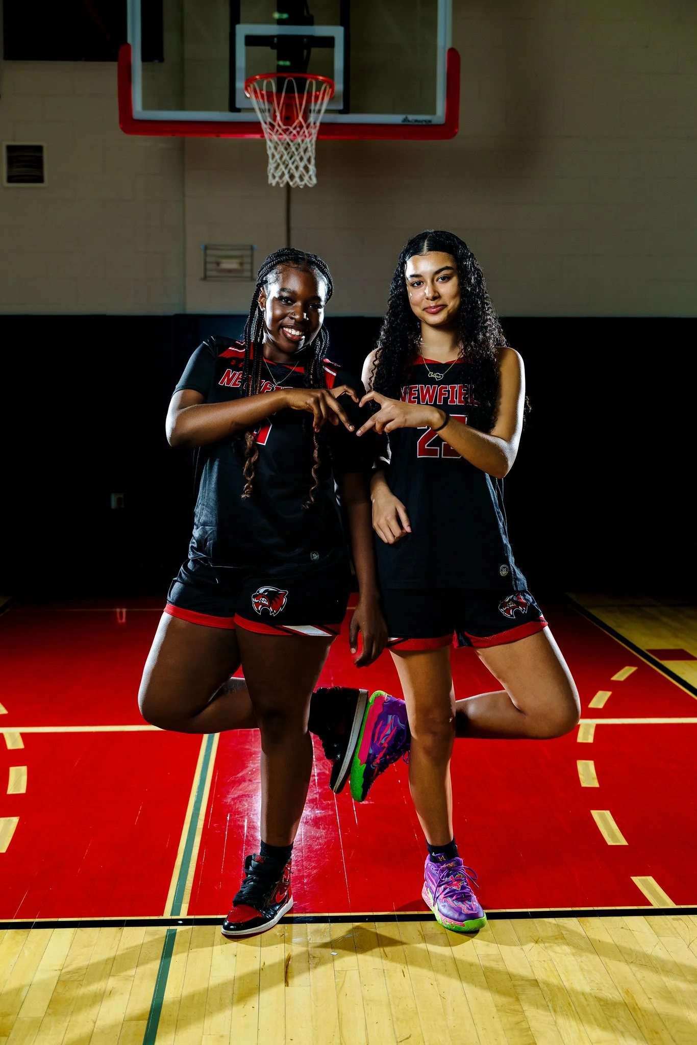 Two female basketball players in black and red uniforms standing on a basketball court, making a heart shape with their hands, with a basketball hoop in the background.