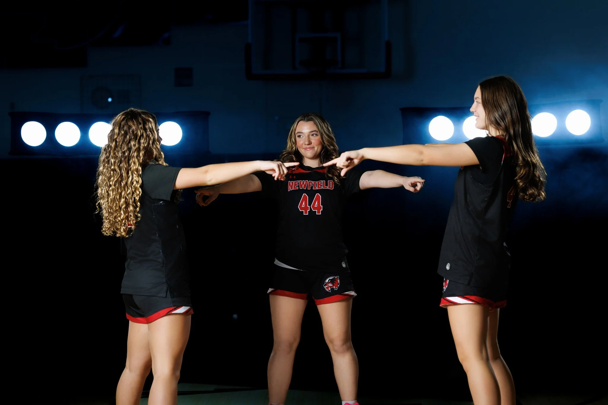Three female volleyball players with matching uniforms standing in a circle on a court, pointing at each other, with dark background and bright gym lights.