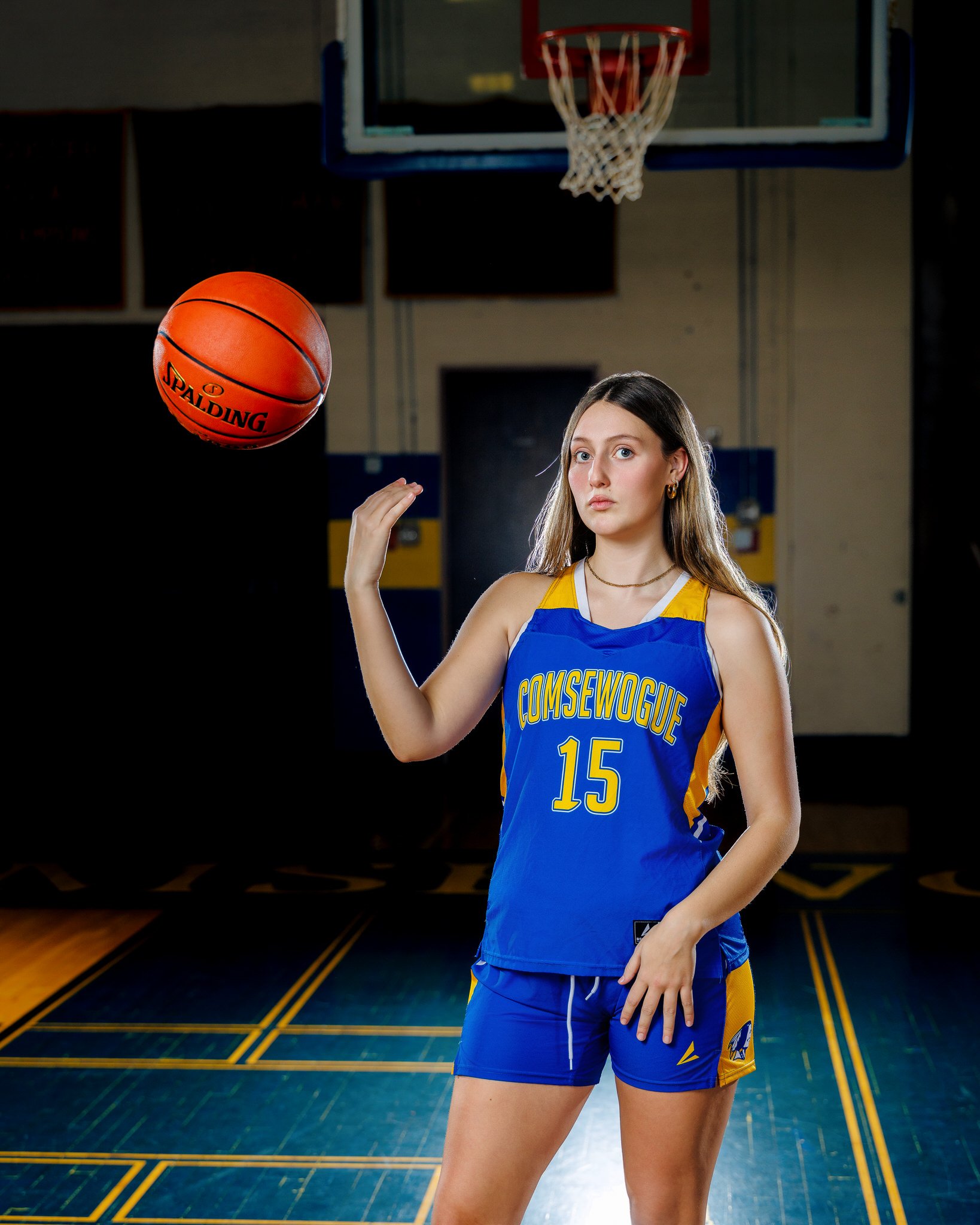 A young woman in a blue and yellow basketball uniform with the number 15 and the name 'COMSEWOGUE' on her jersey, standing on an indoor basketball court with a basketball in the air near her hand.
