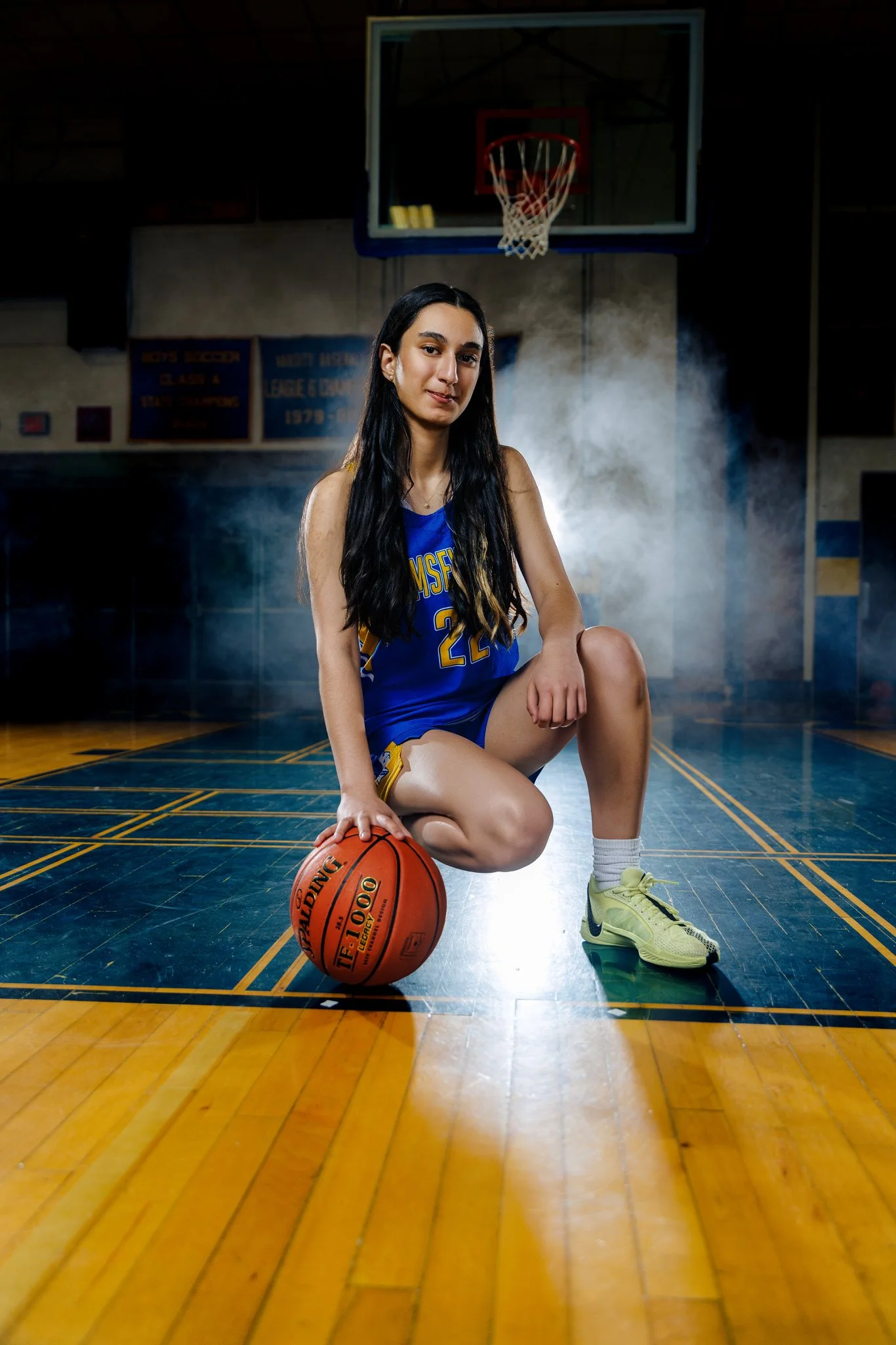 A female basketball player in a blue basketball uniform kneeling on a gymnasium floor with a basketball in her right hand, and a basketball hoop and backboard in the background.