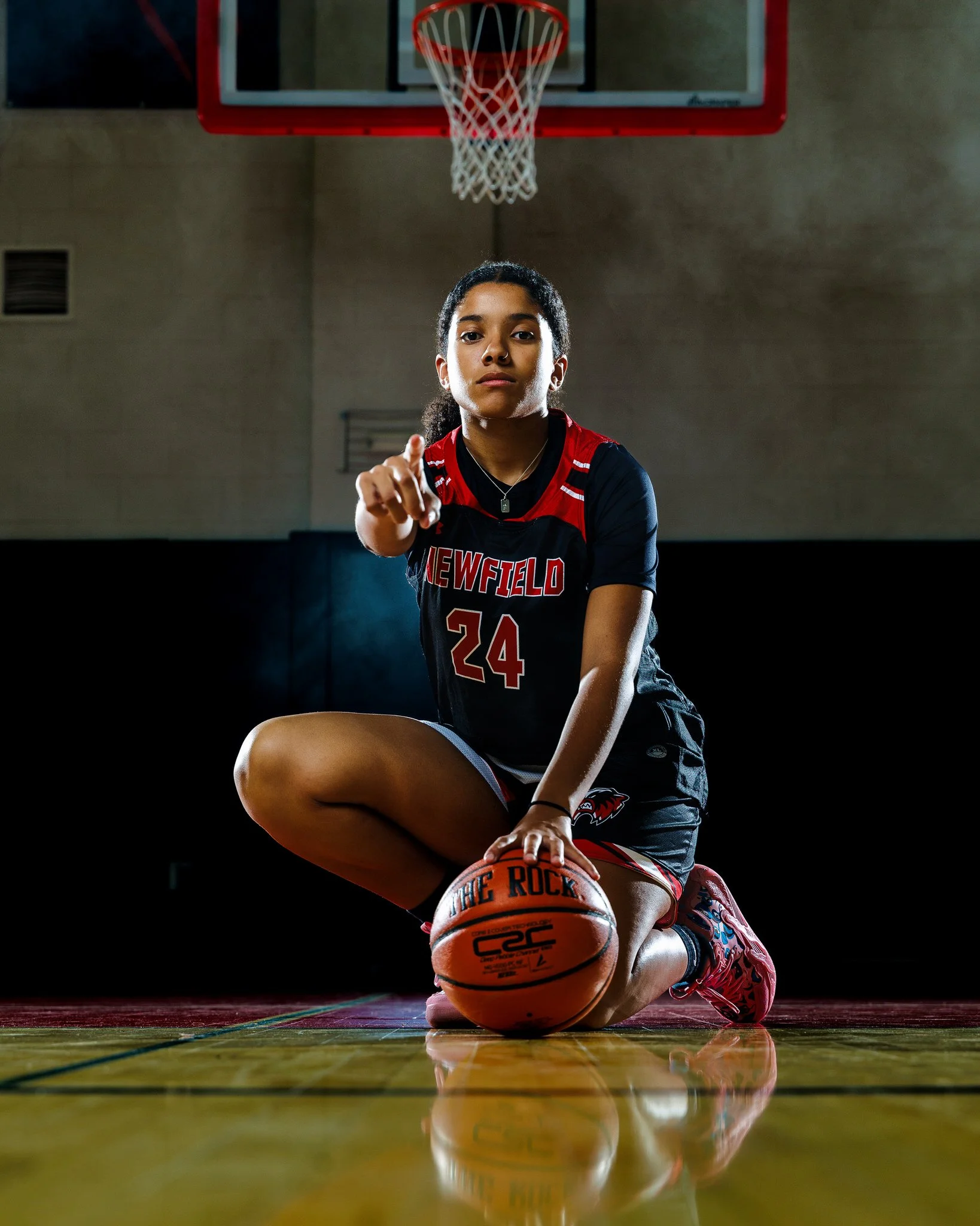 A female basketball player kneeling on the court, holding a basketball, and pointing toward the camera inside a gymnasium with a basketball hoop overhead.