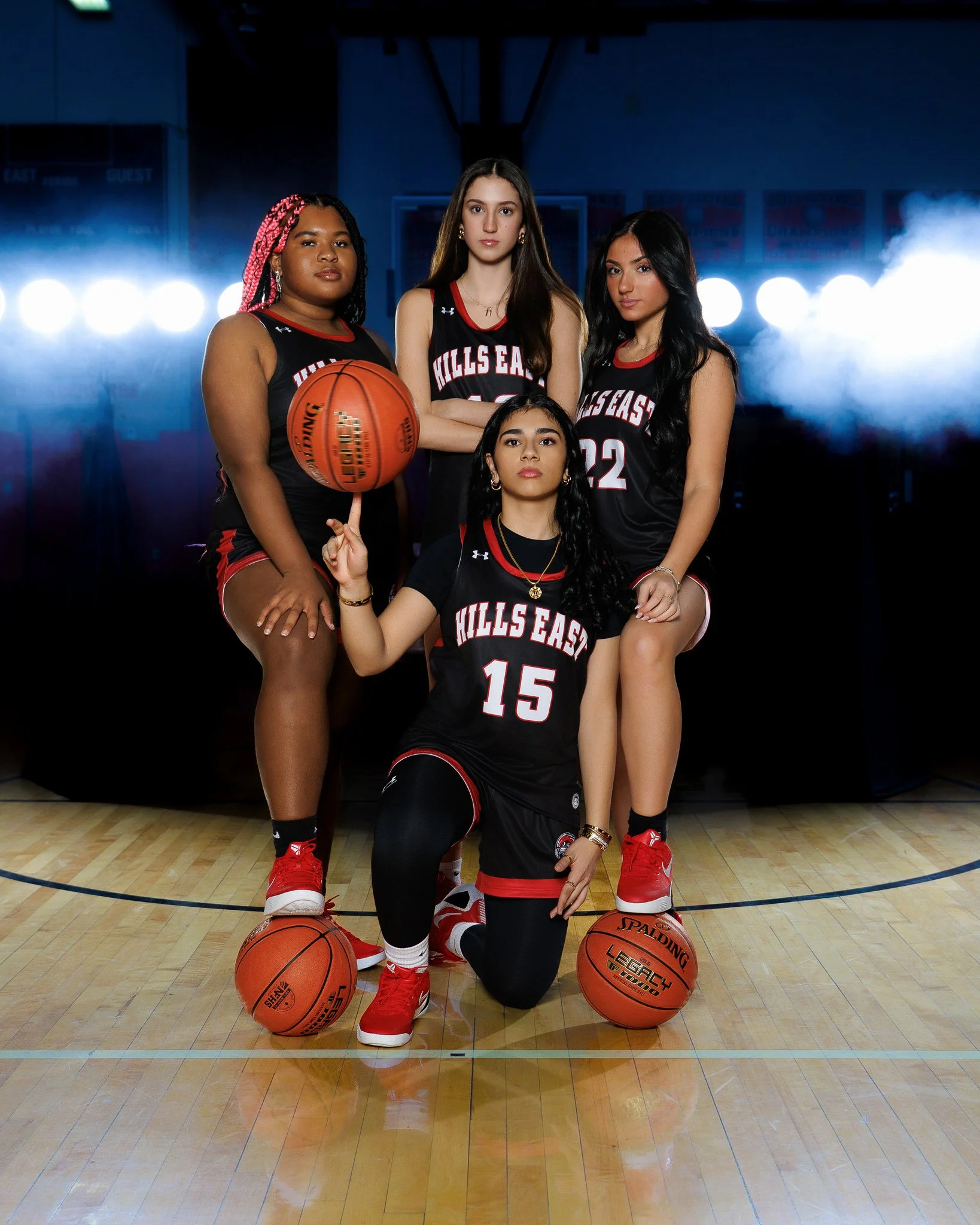 Four young women basketball players in black and red jerseys from Hills East, with two holding basketballs, posing on a gymnasium court with bright lights behind them.