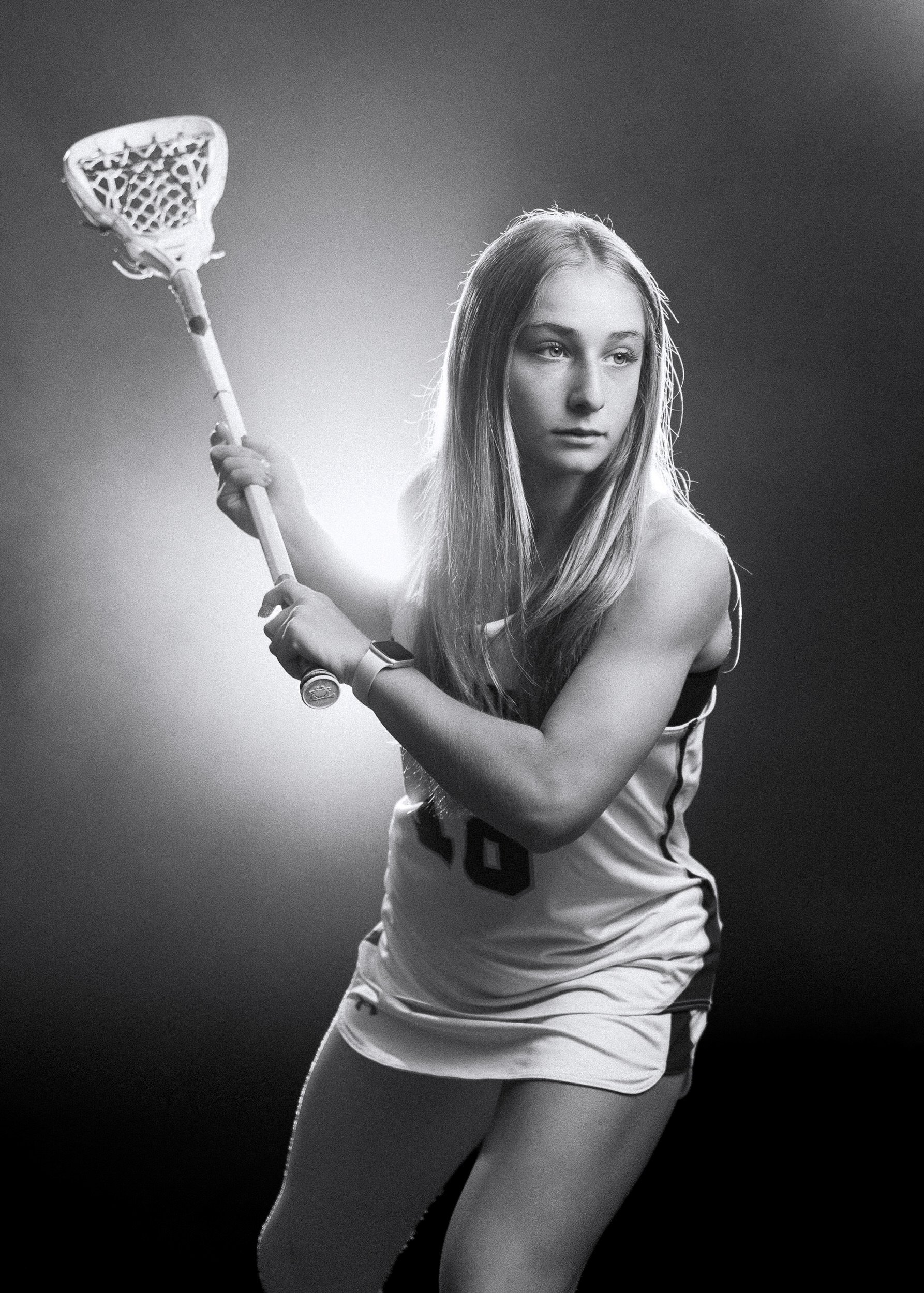 Black and white photo of a female lacrosse player in action with a lacrosse stick, wearing a sports uniform.