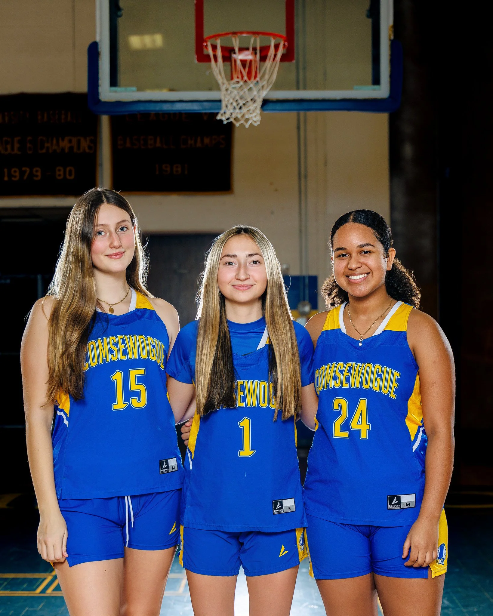 Three young female basketball players standing on a court, wearing blue and yellow uniforms with 'COMSEWOGUE' written on them. They are smiling and standing under a basketball hoop.