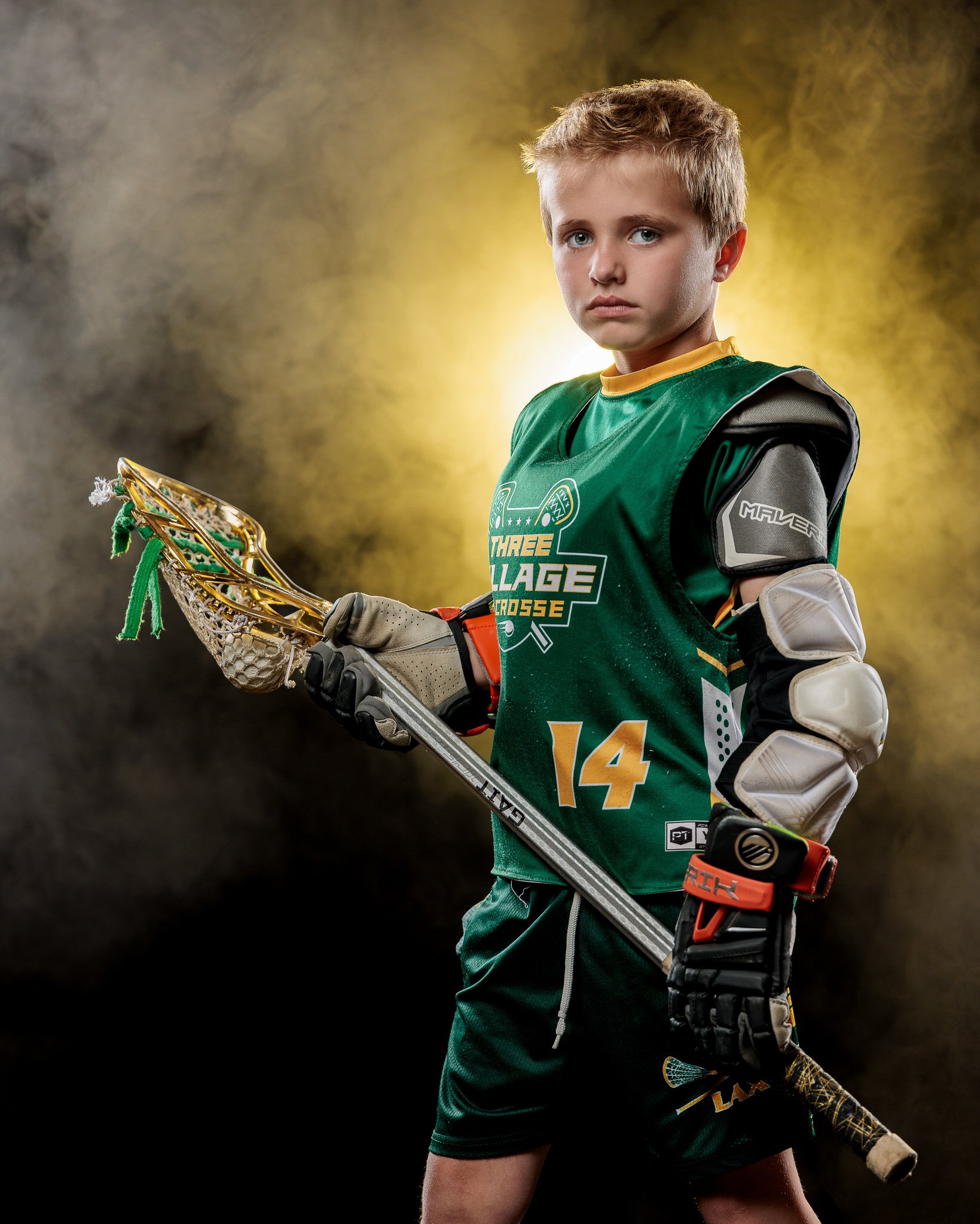 Young lacrosse player in green jersey with number 14 holding a lacrosse stick, wearing protective gear, in front of a smoky background with yellow lighting.
