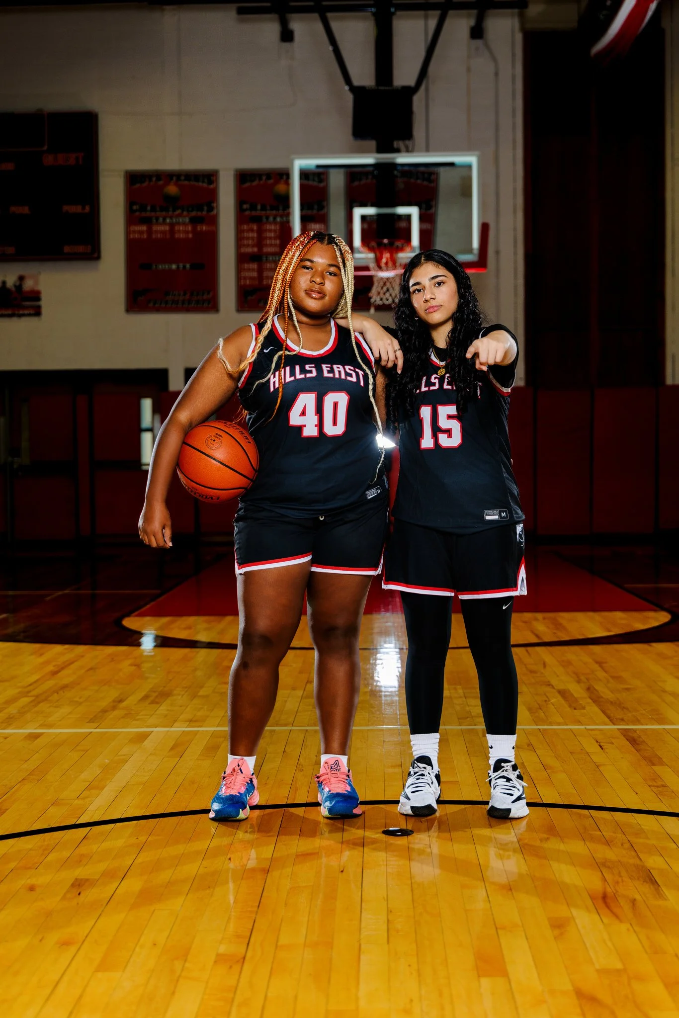 Two female basketball players standing on a basketball court, one holding a basketball, both wearing black jerseys with red and white accents and the words 'Hills East' with their respective numbers, posing confidently.