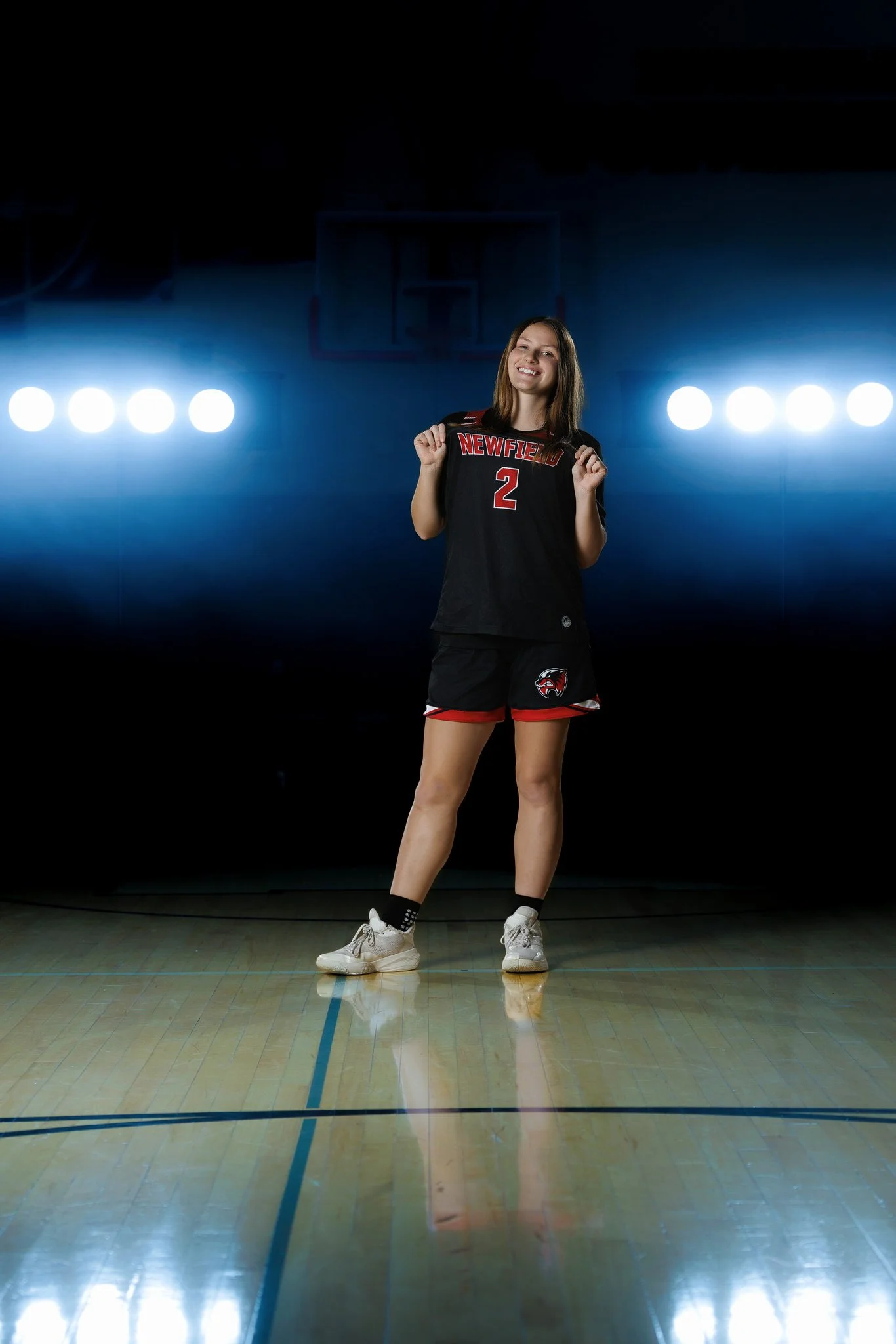 Young female volleyball player standing in a gym on a wooden court, holding a black volleyball jersey with 'NEWFIED 2' and a wolf logo, smiling, illuminated by bright gym lights.