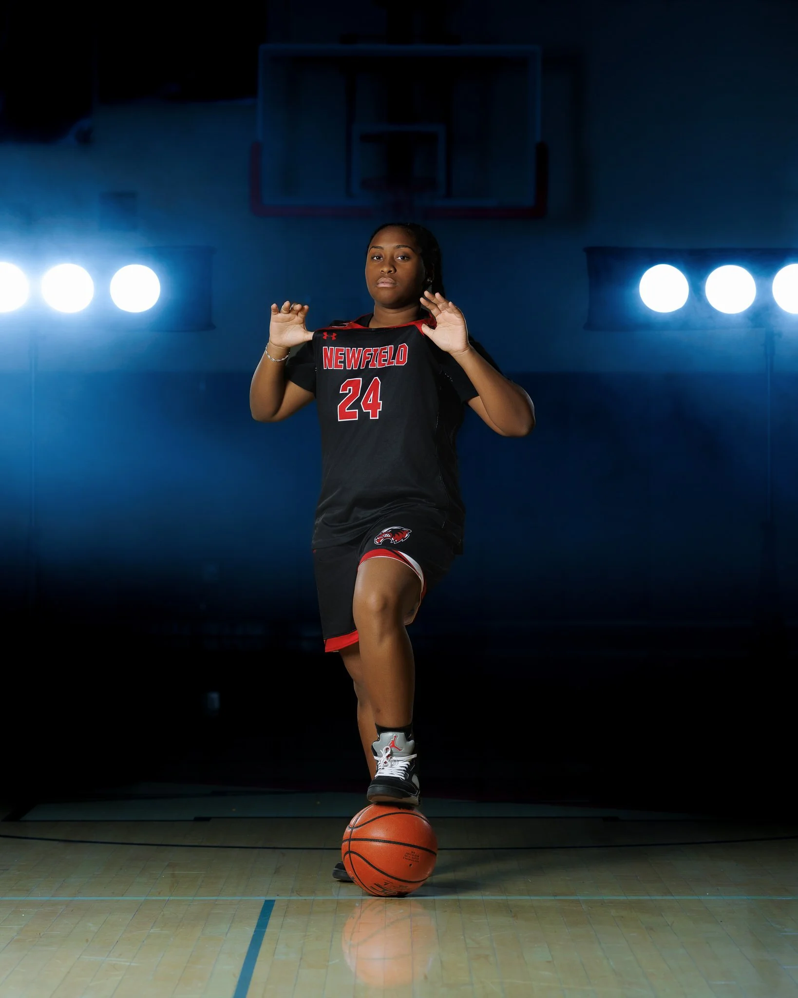 A female basketball player wearing a black and red uniform with the number 24 and 'NEWFELD' on it, balancing a basketball on her foot inside a basketball gymnasium, with bright lights and a basketball hoop in the background.
