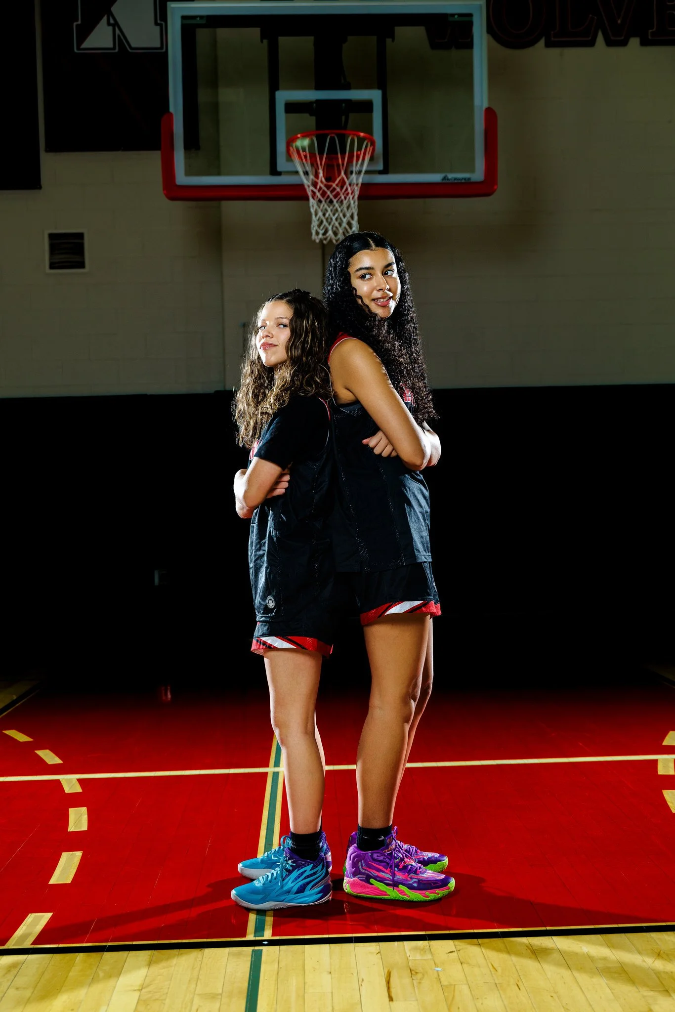 Two female basketball players in athletic gear standing back-to-back on a basketball court, with a basketball hoop in the background.