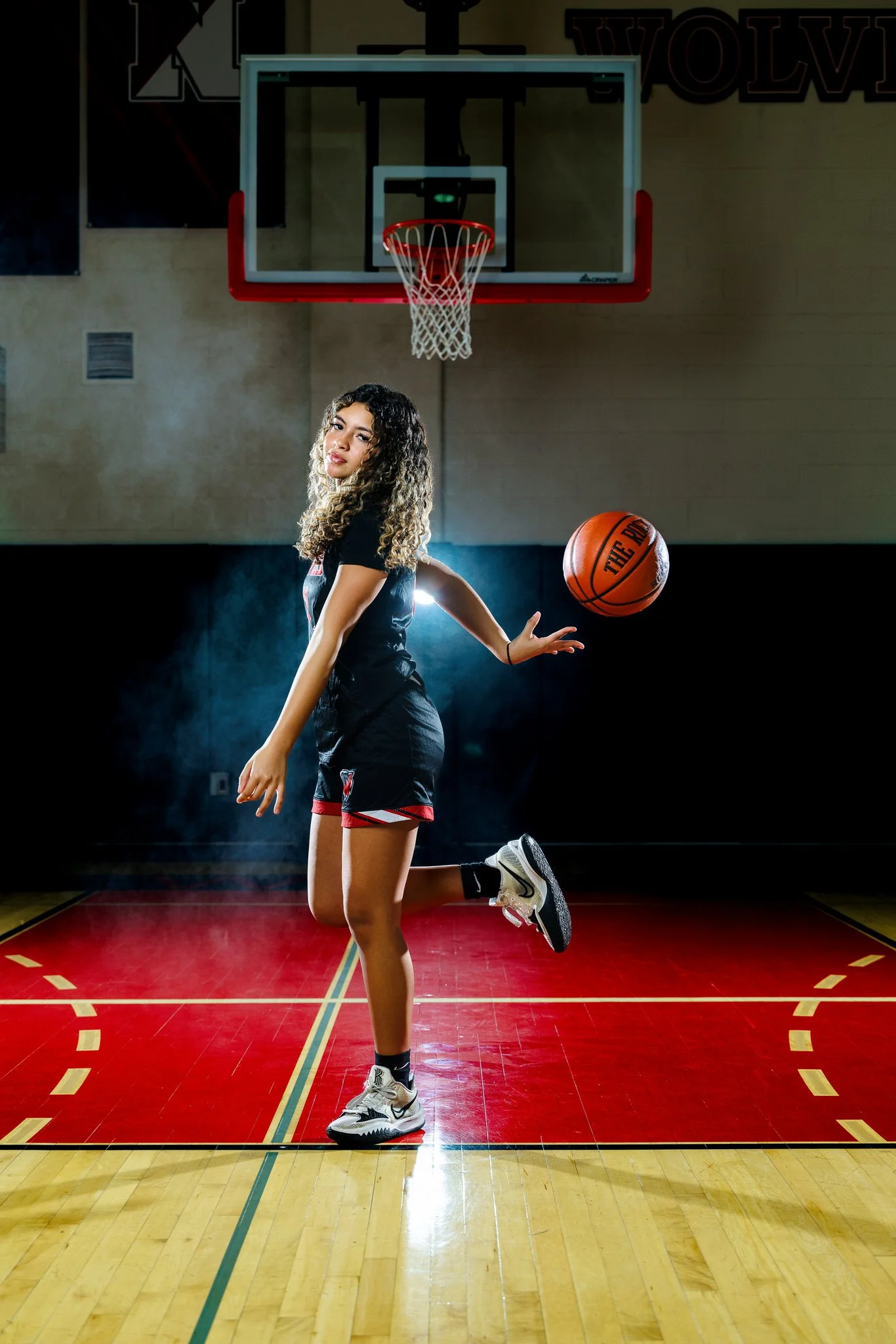 A female basketball player in a black uniform is performing a spin move on a basketball court, with one knee bent and the other leg extended behind her, as a basketball floats nearby.