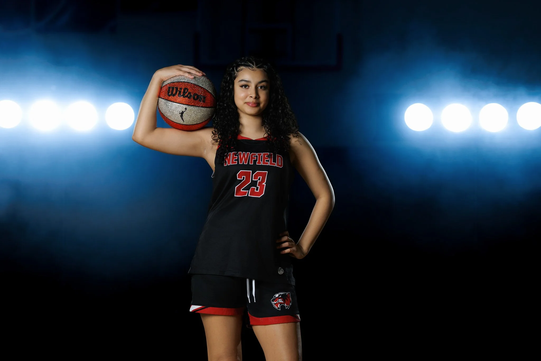 Young female basketball player wearing a black and red Newfield jersey holding a basketball on her right shoulder, standing in a dark gym with bright lights behind her.