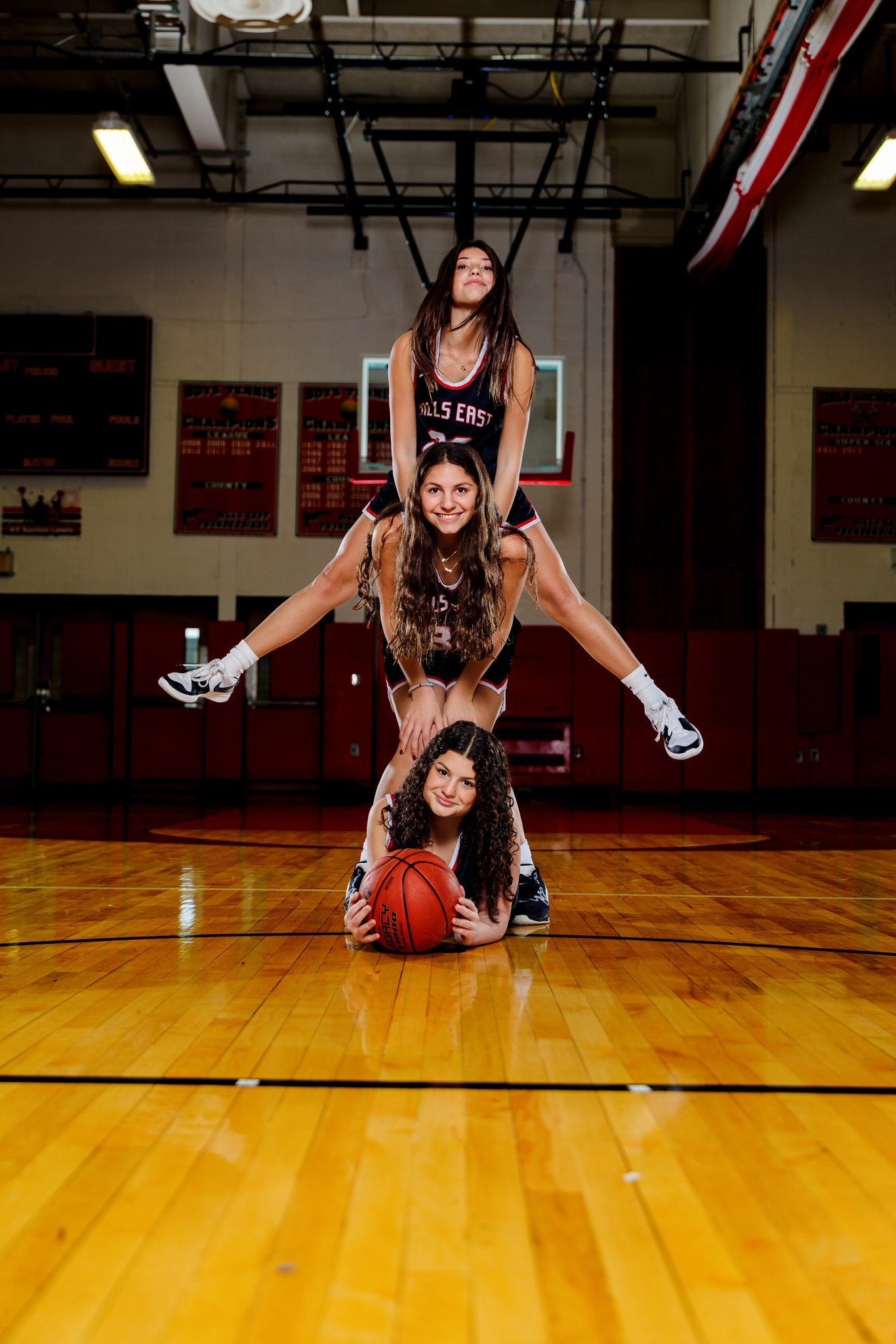 Four young women in basketball uniforms performing a human pyramid on a basketball court inside a gymnasium.