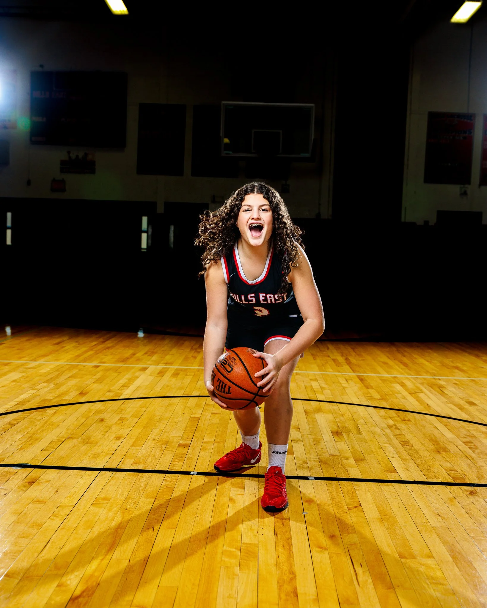 A young girl with curly hair wearing a basketball uniform and red sneakers, holding a basketball on a court, smiling, in a gymnasium.