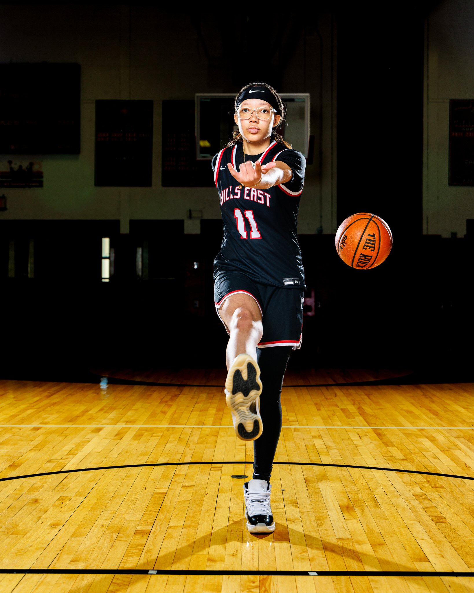 Female basketball player in a black uniform with 'Hills East' and the number 11, practicing on an indoor court. She is wearing glasses, a headband, and reaching towards the camera while dribbling a basketball.