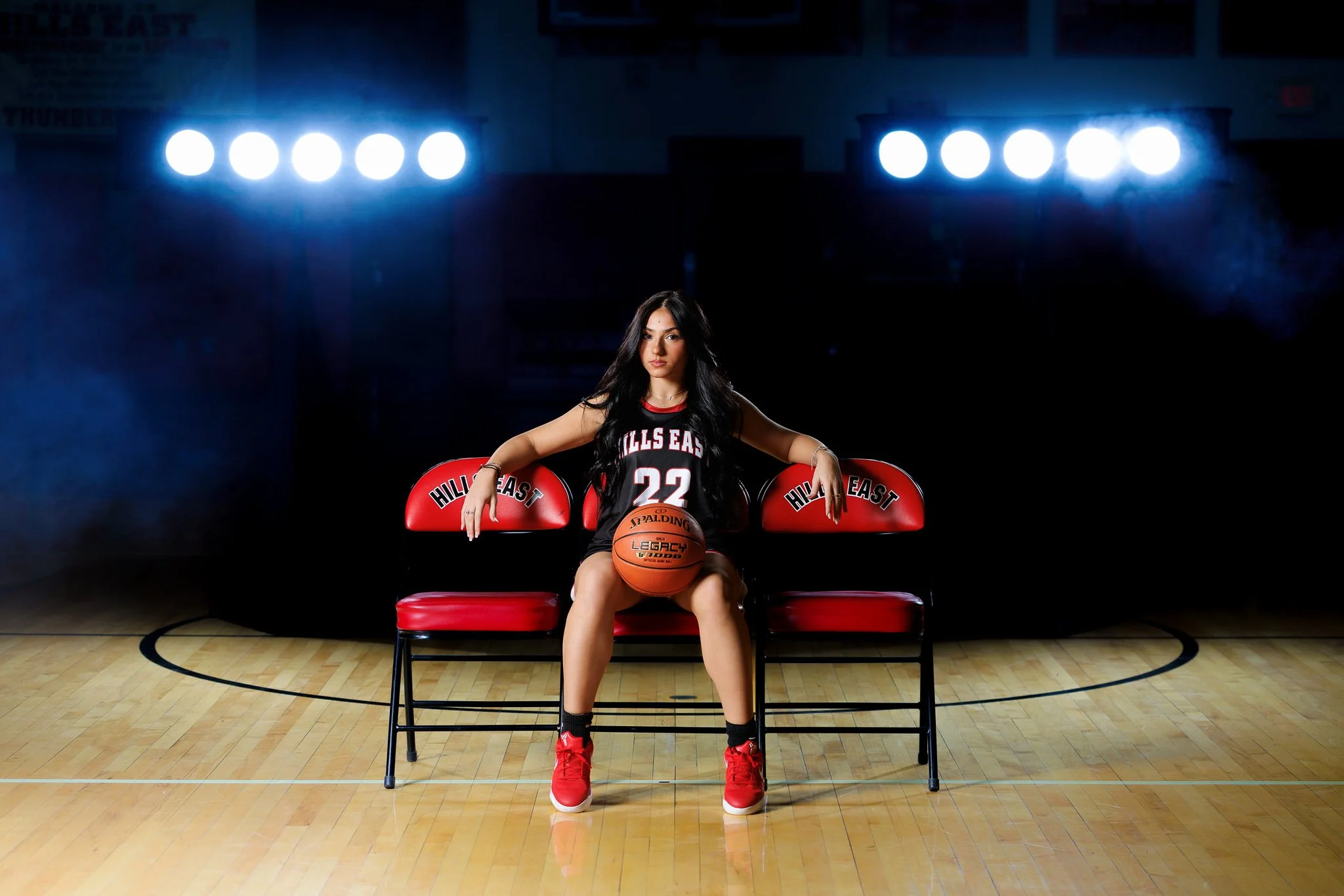 A young female basketball player sitting on a bench on a basketball court, holding a basketball between her legs, wearing a black jersey with the number 22, red shoes, and black socks, with bright lights overhead.