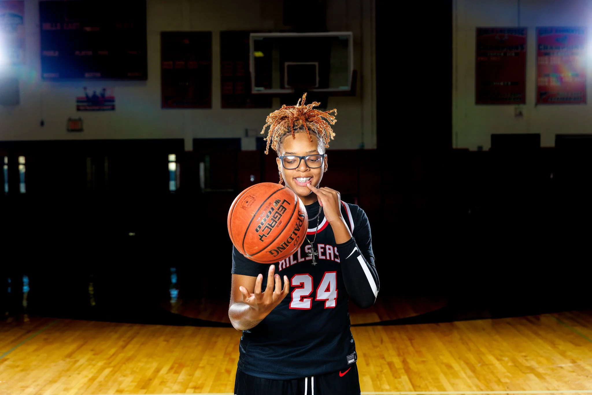 A young woman with blonde dreadlocks and glasses in a basketball gym, holding a basketball and smiling.