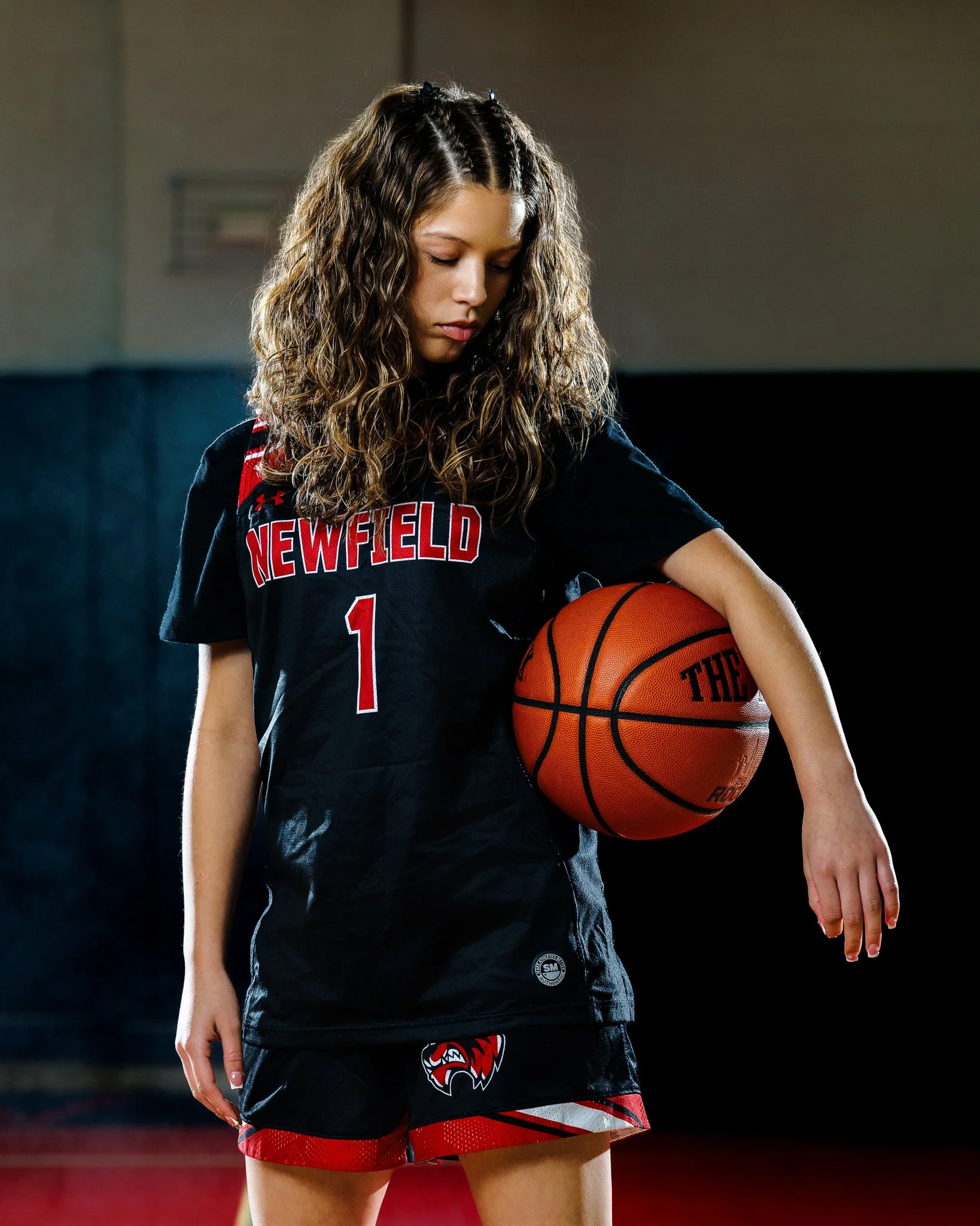 A young female basketball player with curly hair stands holding a basketball against a dark gym background. She wears a black sports jersey with red and white lettering and shorts with red and black accents.